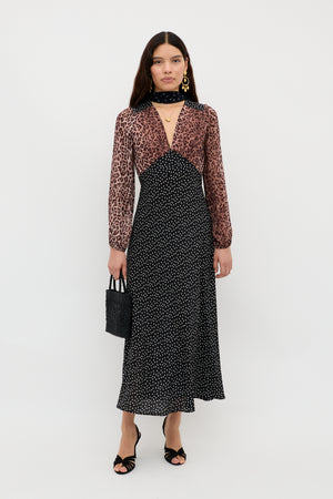 A woman stands against a white background wearing the Kaila - Leopard Spot Brown dress, featuring a sheer leopard print top with black polka-dot skirt. She accessorizes with a small black handbag, gold jewelry, sandals, and a scarf.