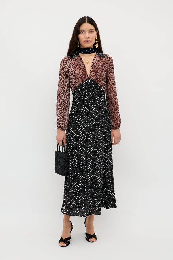 A woman stands against a white background wearing the Kaila - Leopard Spot Brown dress, featuring a sheer leopard print top with black polka-dot skirt. She accessorizes with a small black handbag, gold jewelry, sandals, and a scarf.