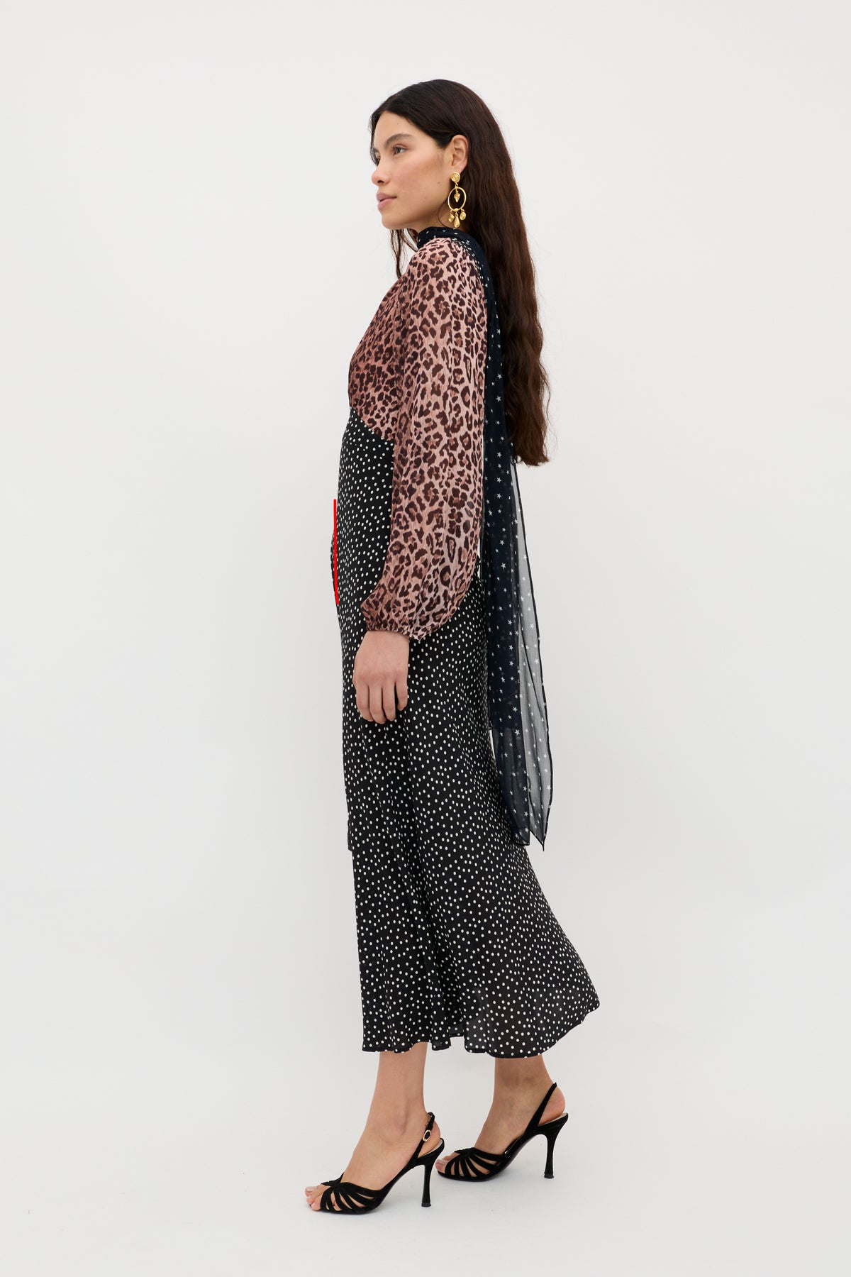 A woman stands in profile against a white background, wearing the Kaila - Leopard Spot Brown dress with sheer leopard-print sleeves, a polka dot skirt, and strappy black high-heeled sandals.