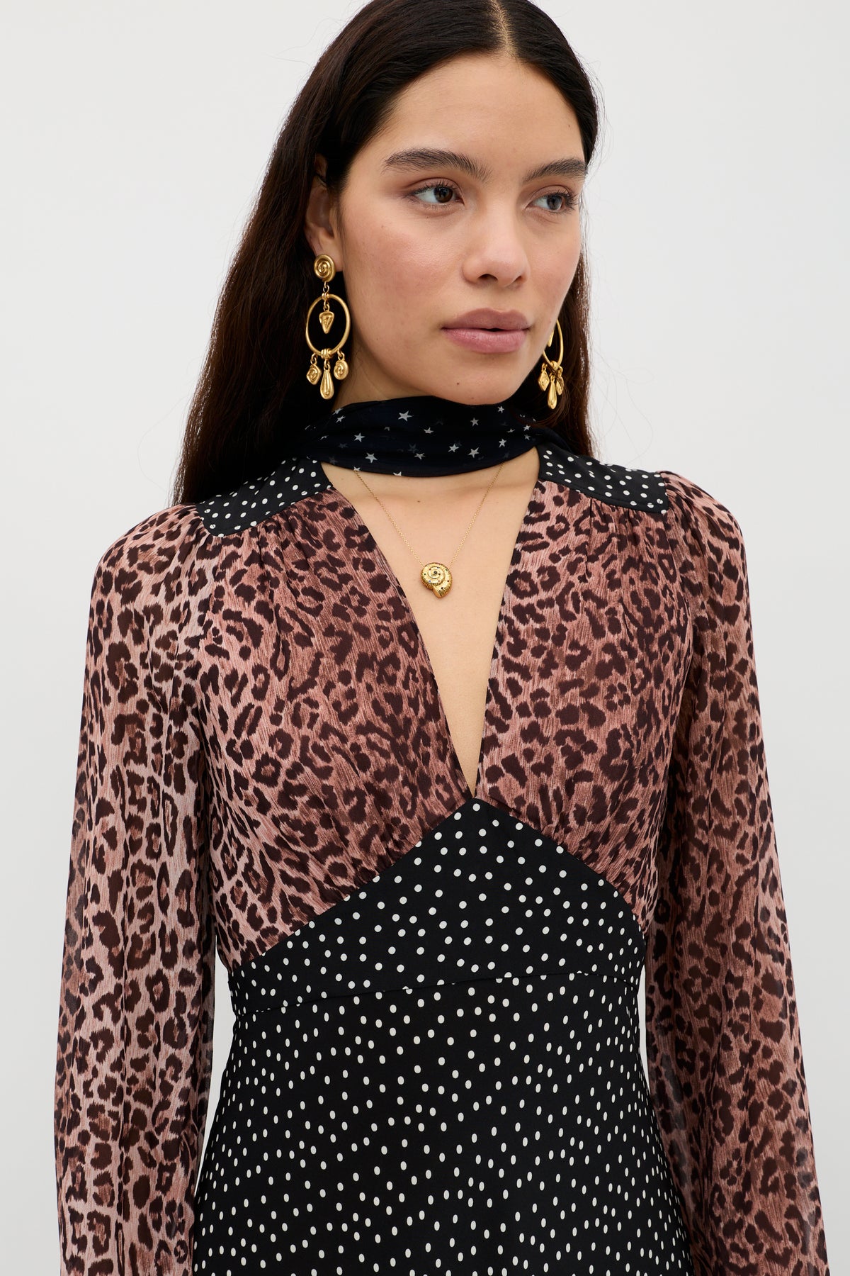 A woman with long dark hair wears the Kaila - Leopard Spot Brown dress, featuring a flattering vintage shape with leopard print on top and polka dots below. She adds gold earrings, layered necklaces, and a black scarf with white stars.