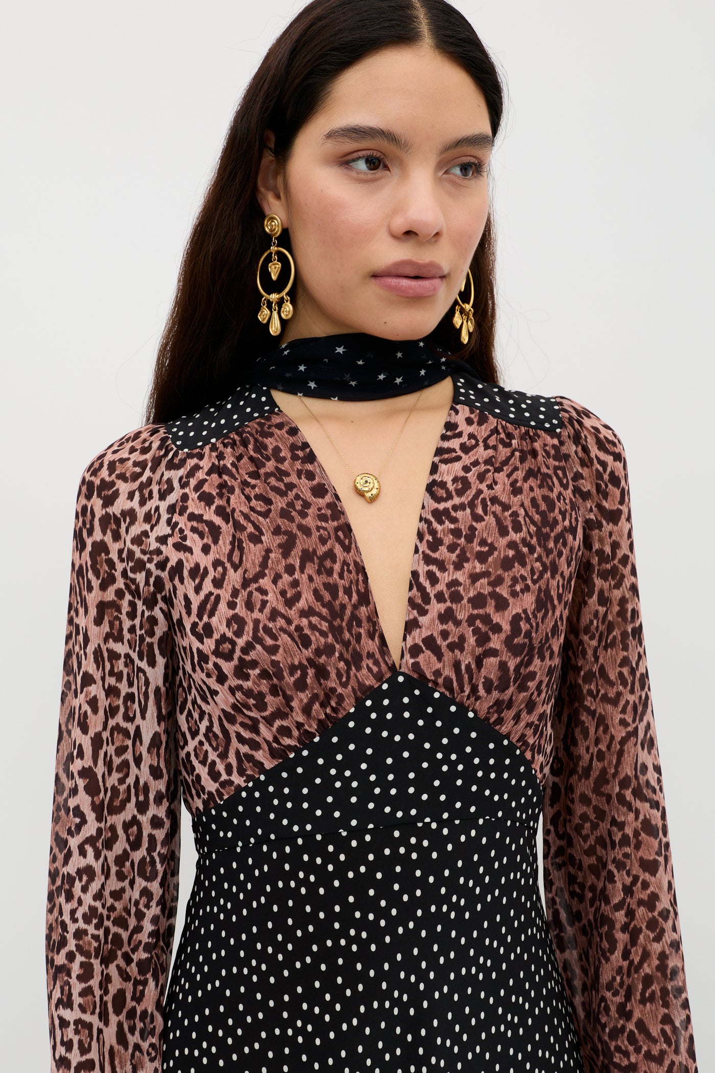 A woman with long dark hair wears the Kaila - Leopard Spot Brown dress, featuring a flattering vintage shape with leopard print on top and polka dots below. She adds gold earrings, layered necklaces, and a black scarf with white stars.