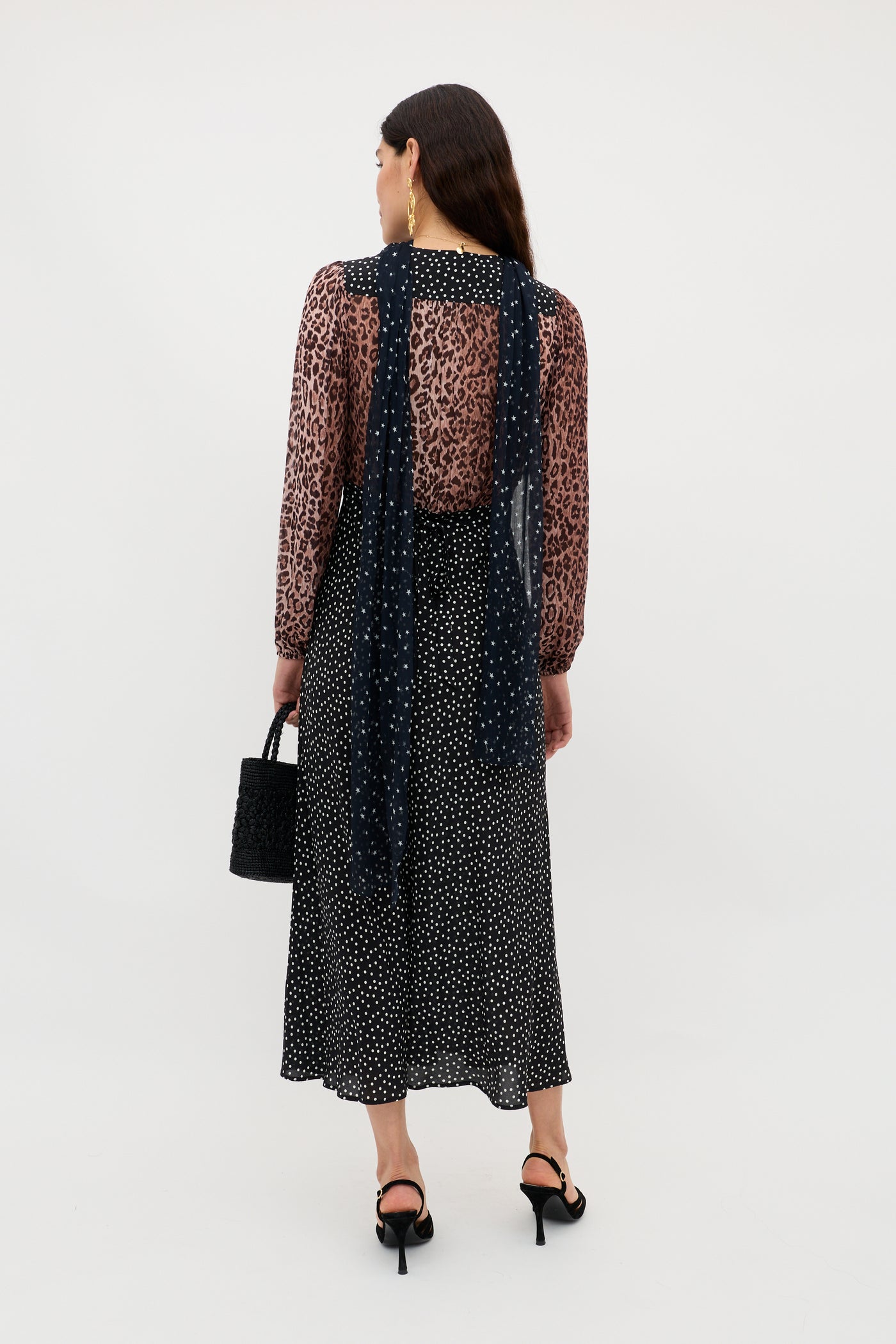 A woman with loose long dark hair faces away, wearing the Kaila - Leopard Spot Brown sheer long-sleeve top, a black and white polka dot midi skirt, black heeled sandals, and holding a black handbag.