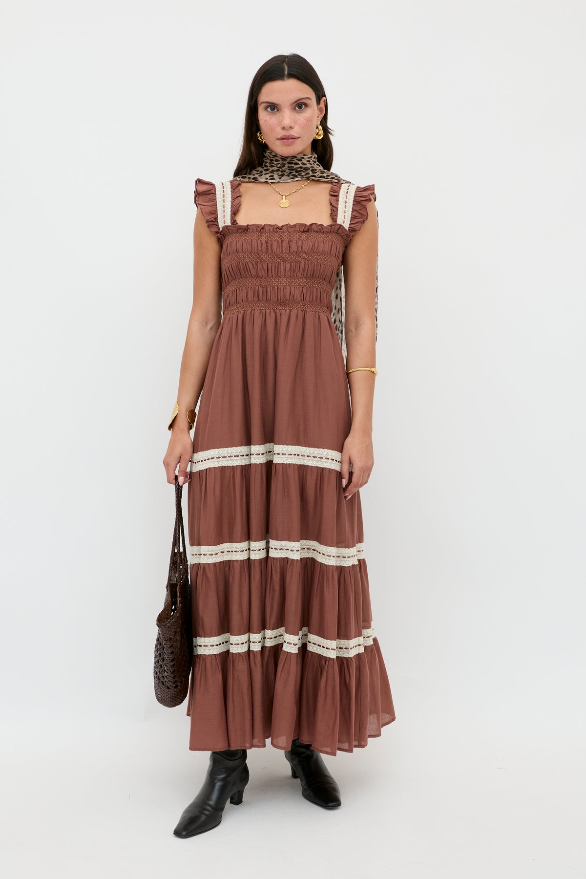 A woman with straight brown hair and gold jewelry stands against a white background wearing the vintage-inspired Aubrie - Brown dress with ruffled lace trim, styled with black boots, a patterned scarf, and a woven handbag for an effortlessly chic look.