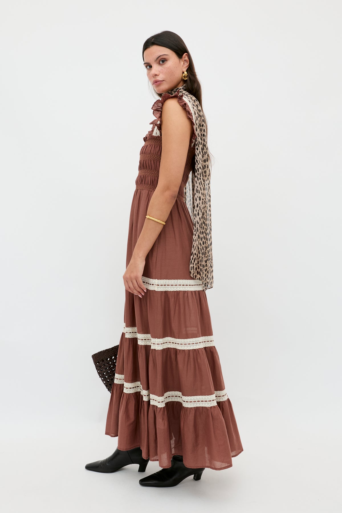 A woman stands sideways in the Aubrie - Brown dress with cream lace crochet midi accents, paired with black ankle boots, gold jewelry, a leopard-print scarf, and a dark woven bag against a plain white background.