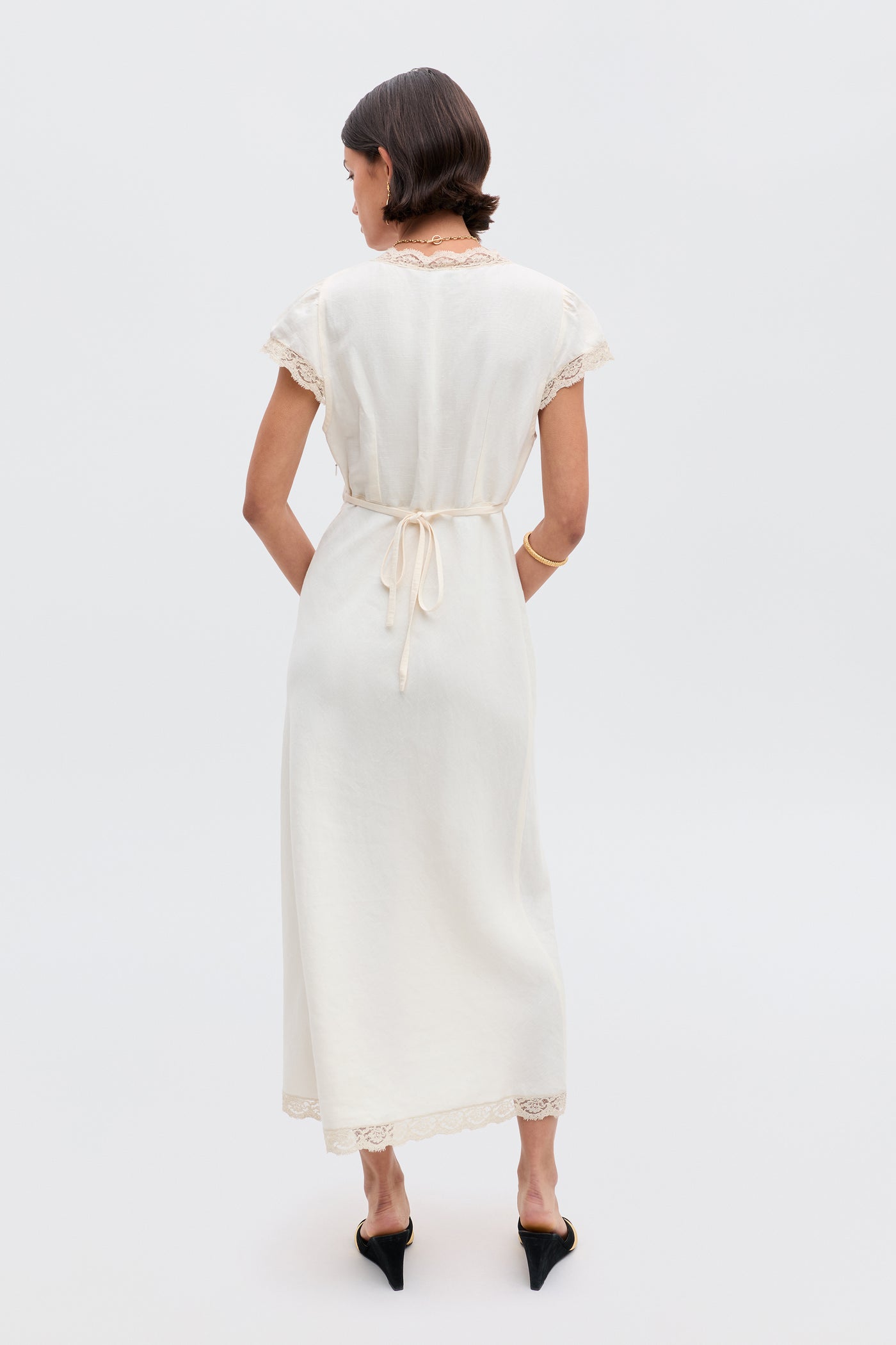 A woman stands facing away in the Clarice - Cream vintage nightdress with lace trim, a waist tie, and black heels. She has chin-length straight hair and is posed against a plain, light-colored background.