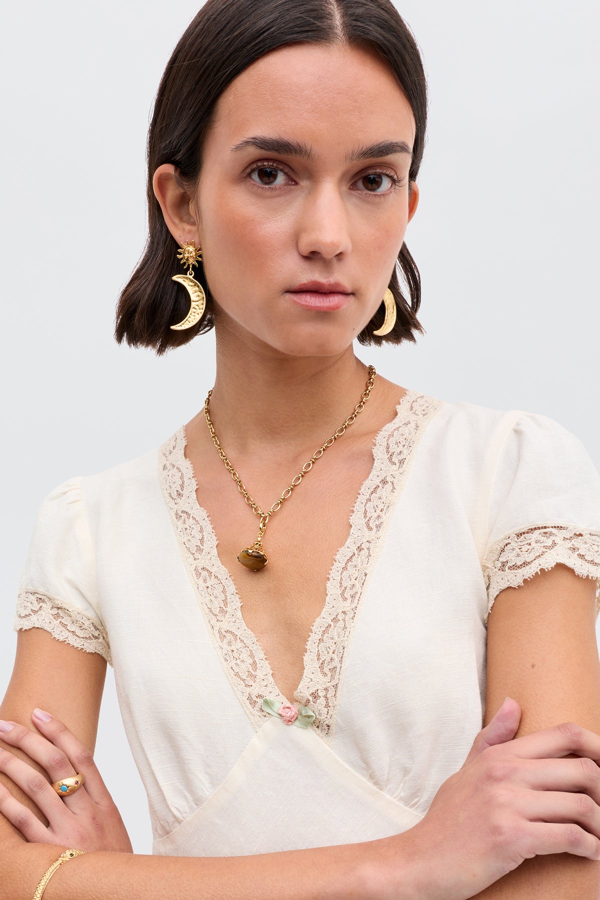 A woman with short dark hair wears the Clarice - Cream vintage nightdress with lace trim, plus a gold moon earring, heart pendant necklace, and ring. She stands with arms crossed against a plain background.