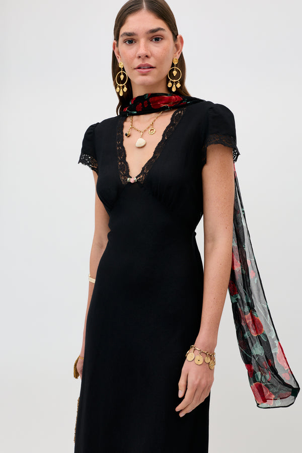 A woman stands against a plain light background, dressed in a black dress with lace trim and gold jewelry, with the Julien - Claudia Rose Black scarf—featuring red and green patterns—draped around her neck.