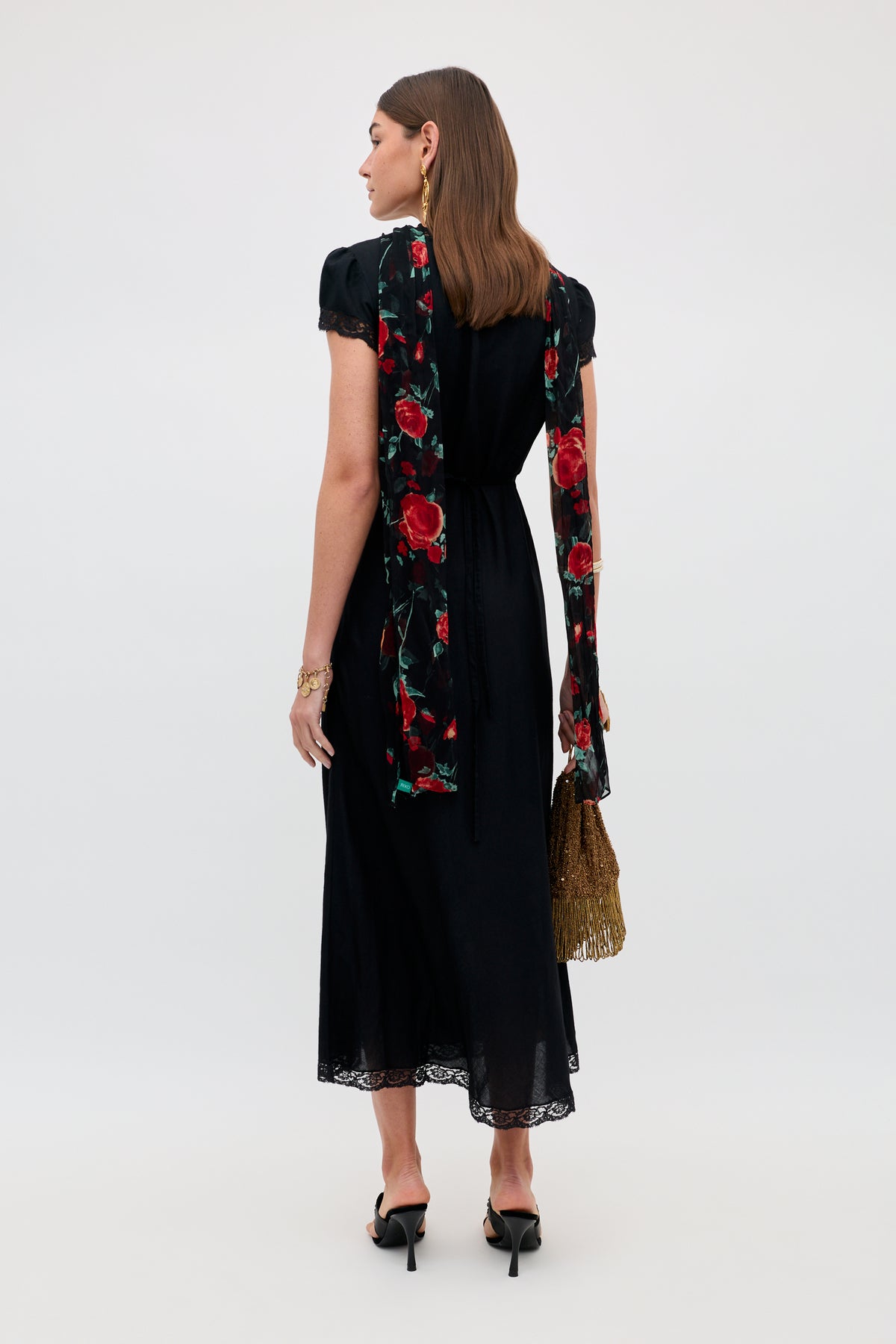 A woman faces away, wearing the Clarice - Black midi dress with lace trim and a long red floral scarf. She holds a gold fringe handbag, wears black heels, has straight brown hair styled neatly, and earrings.