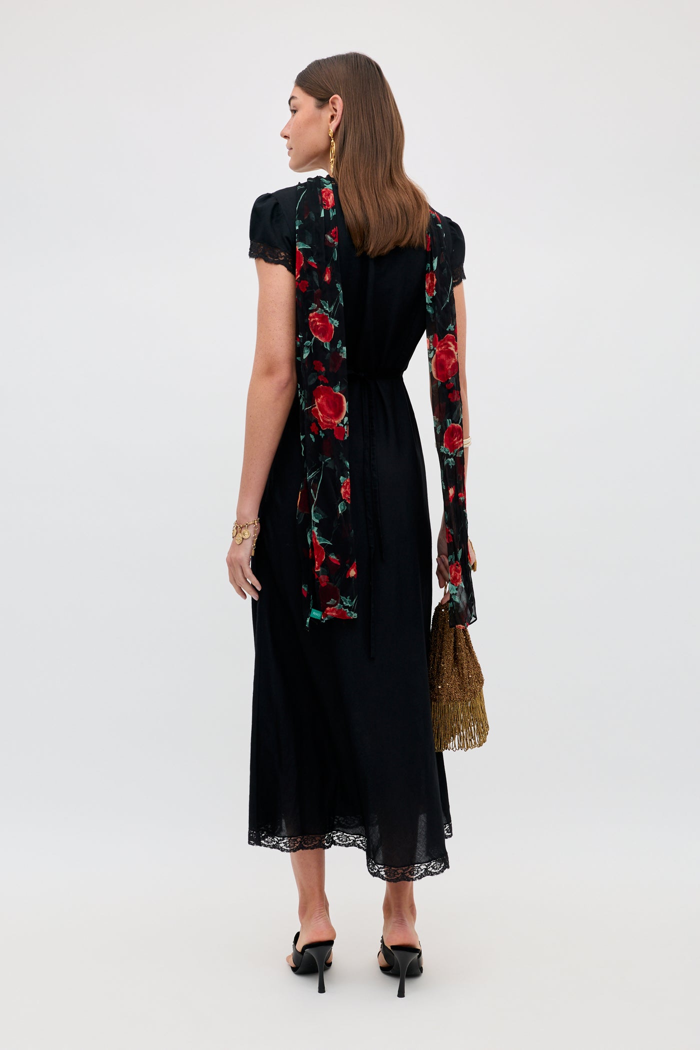 A woman faces away, wearing the Clarice - Black midi dress with lace trim and a long red floral scarf. She holds a gold fringe handbag, wears black heels, has straight brown hair styled neatly, and earrings.