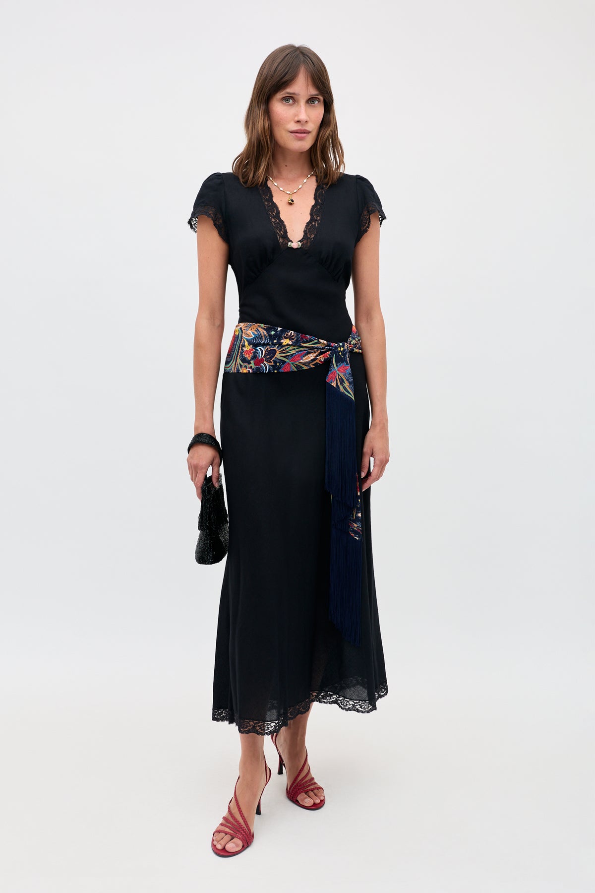 Wearing the Clarice - Black dress with lace trim, a vibrant embroidered sash with fringe, red strappy heels, layered necklaces, and holding a small black handbag, a woman stands against a plain light background.