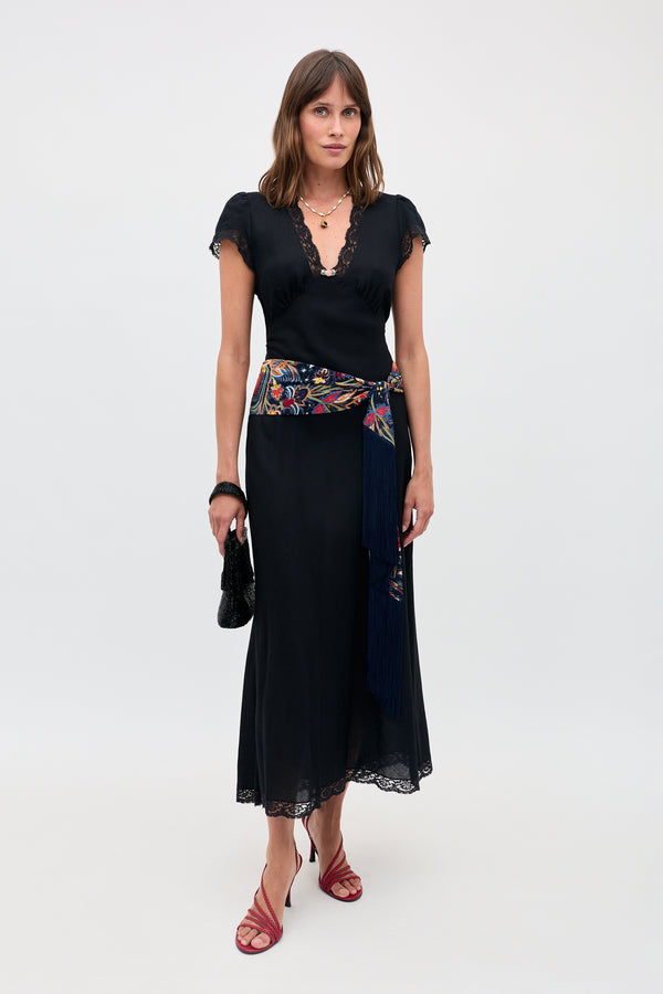 Wearing the Clarice - Black dress with lace trim, a vibrant embroidered sash with fringe, red strappy heels, layered necklaces, and holding a small black handbag, a woman stands against a plain light background.