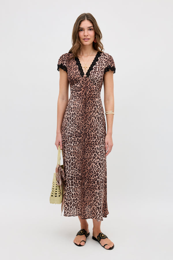 A woman stands against a white background wearing the Clarice - Brown Leopard short-sleeve, V-neck midi dress with black lace trim. She accessorizes with a woven bag, black sandals, and gold jewelry.