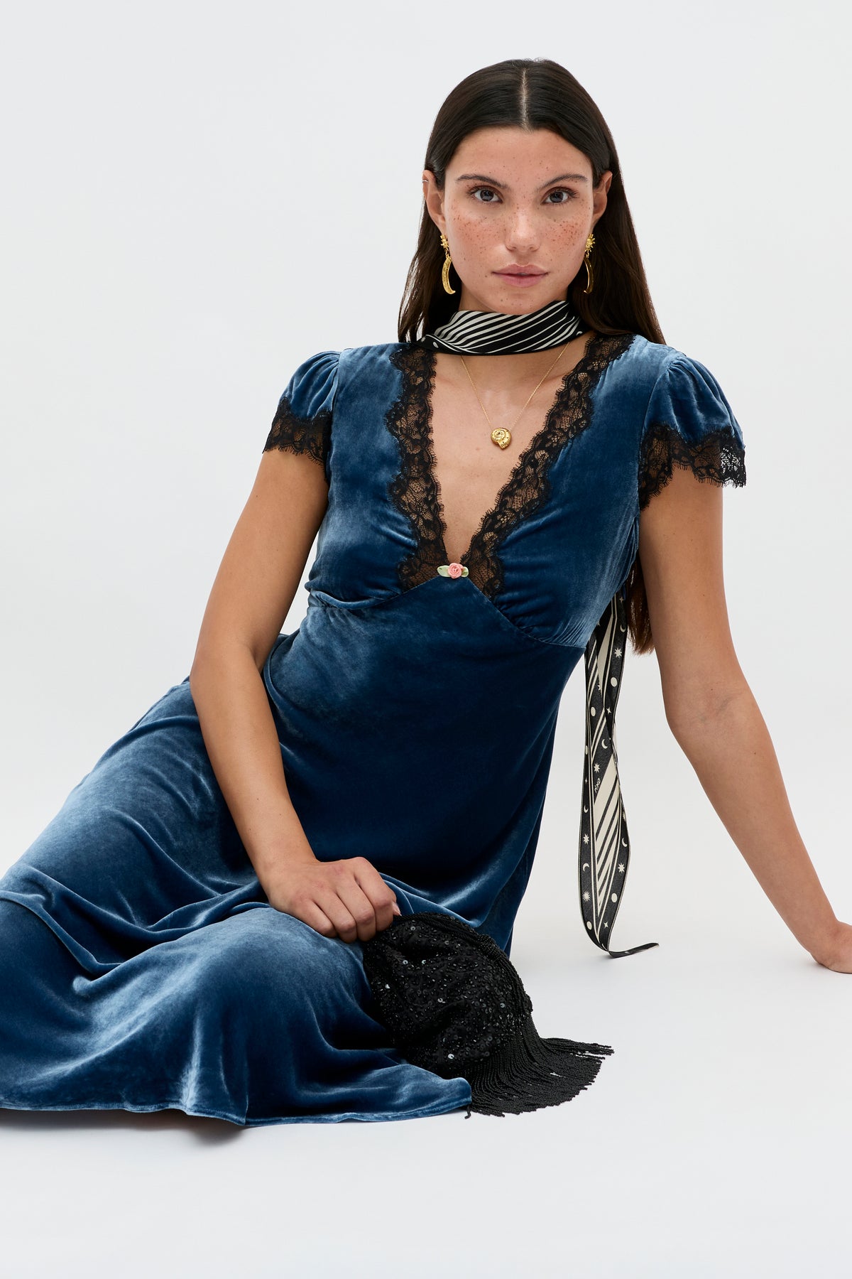 A woman with long dark hair sits on the floor wearing a deep blue velvet dress, gold jewelry, and holds the Clarice - Steel Blue clutch bag.