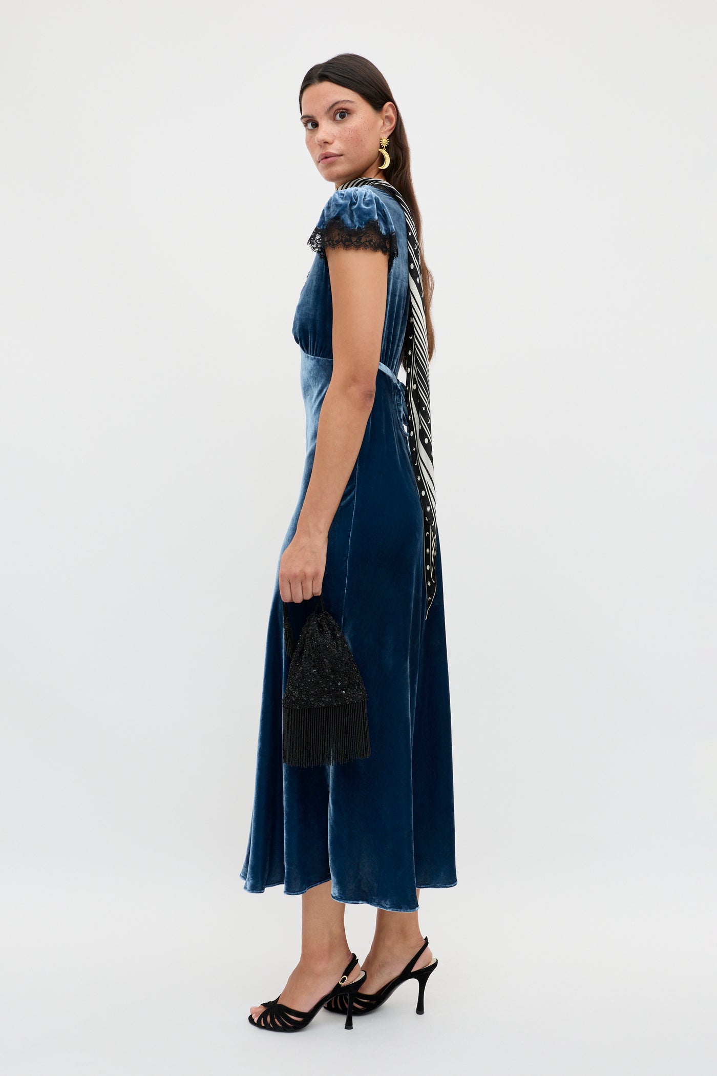 A woman wears the Clarice - Steel Blue velvet midi dress with lace sleeves, black heels, gold earrings, and holds a black fringed handbag. She stands sideways with long brown hair against a plain white background.