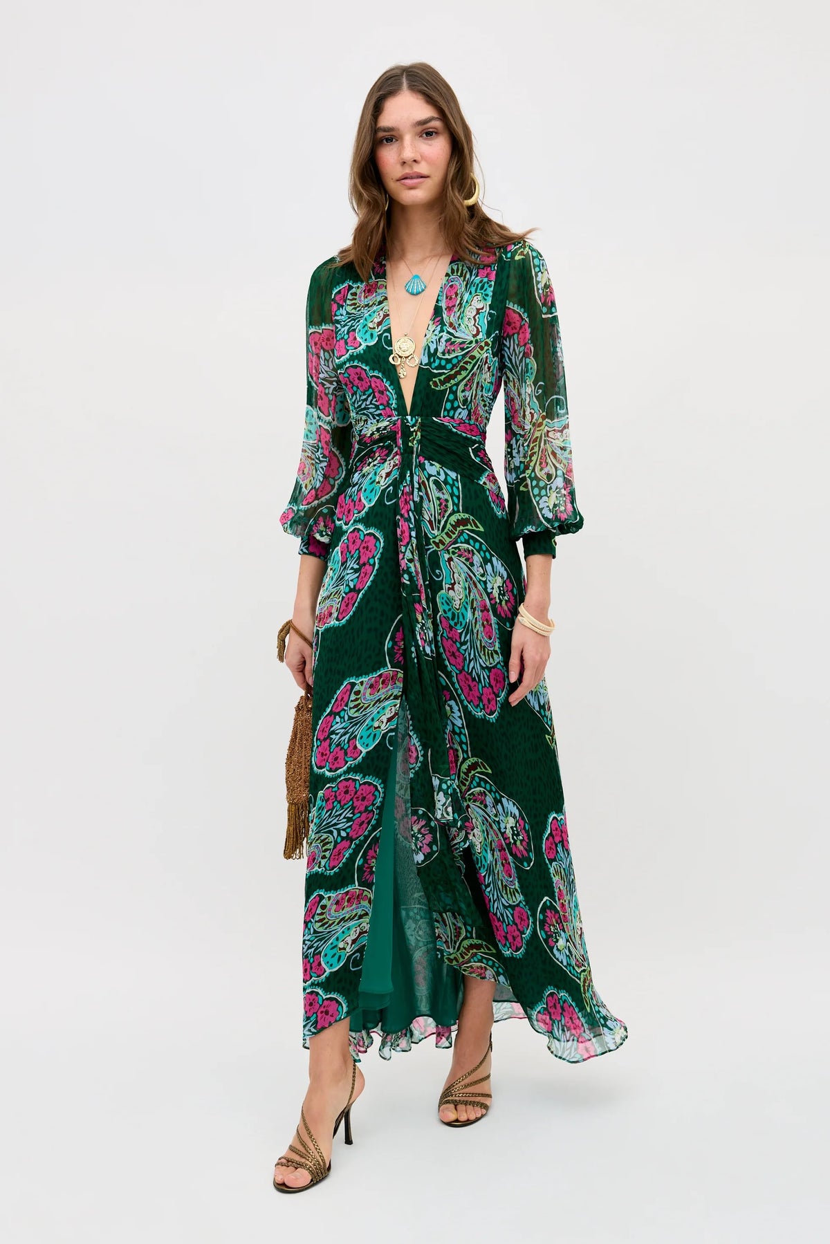 Wearing the Meera - Butterfly Bloom Green dress, a woman stands against a white background. The long green dress features a patterned design, deep V-neck, and three-quarter sleeves. She accessorizes with gold jewelry, sandals, and a brown fringe clutch.