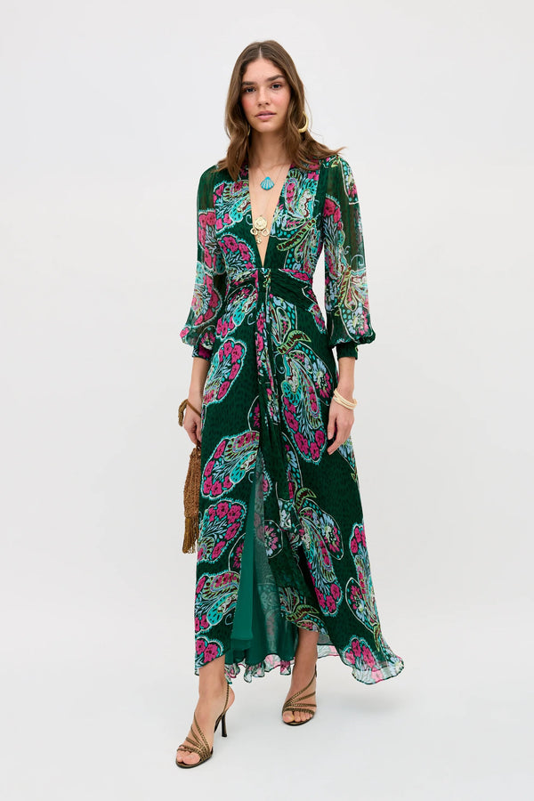 Wearing the Meera - Butterfly Bloom Green dress, a woman stands against a white background. The long green dress features a patterned design, deep V-neck, and three-quarter sleeves. She accessorizes with gold jewelry, sandals, and a brown fringe clutch.