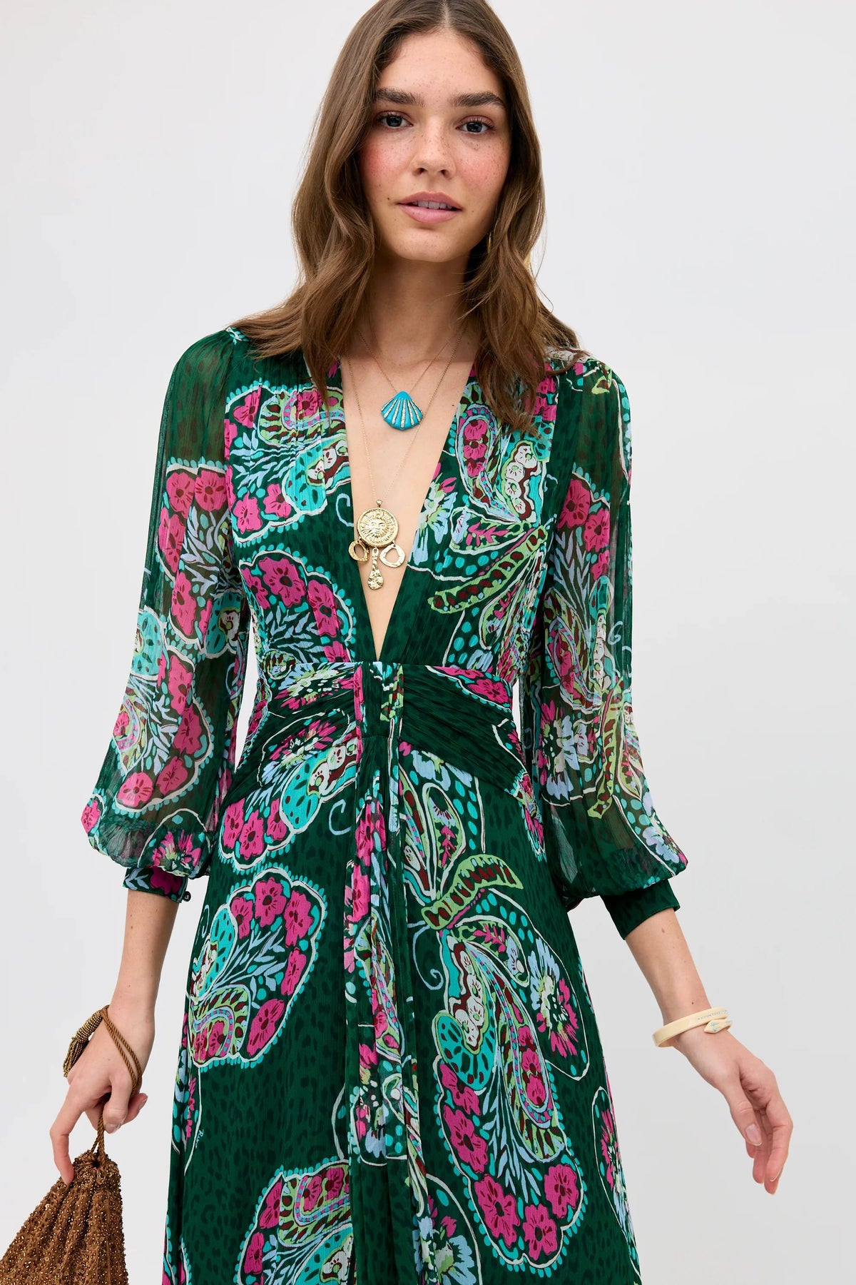 A woman wearing the Meera - Butterfly Bloom Green dress with a colorful paisley pattern, layered necklaces, and holding a brown textured bag stands against a plain light background.