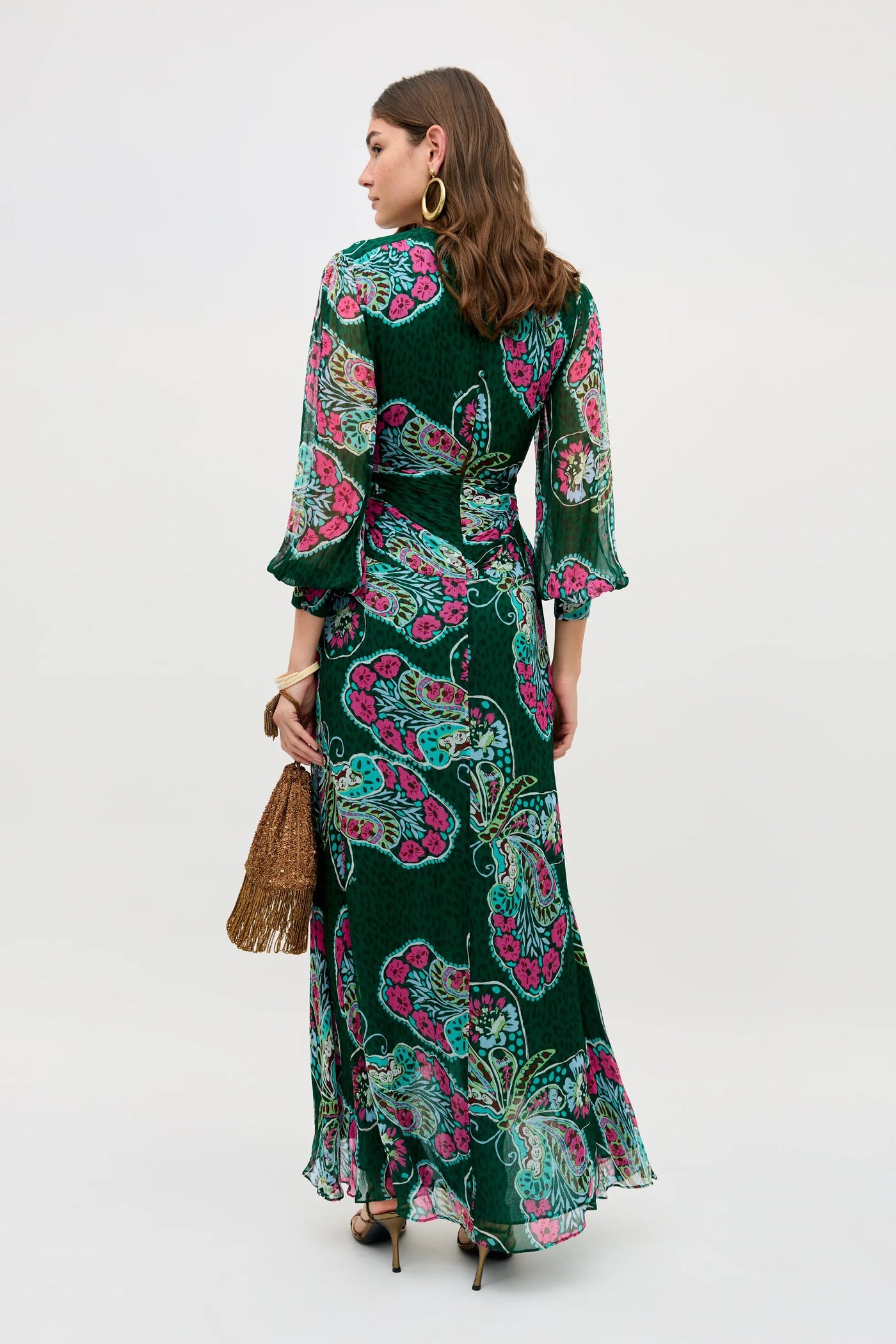 A woman faces away, wearing the Meera - Butterfly Bloom Green dress with pink and teal florals. She carries a brown fringed handbag, wears hoop earrings, and her wavy brown hair falls to her shoulders.