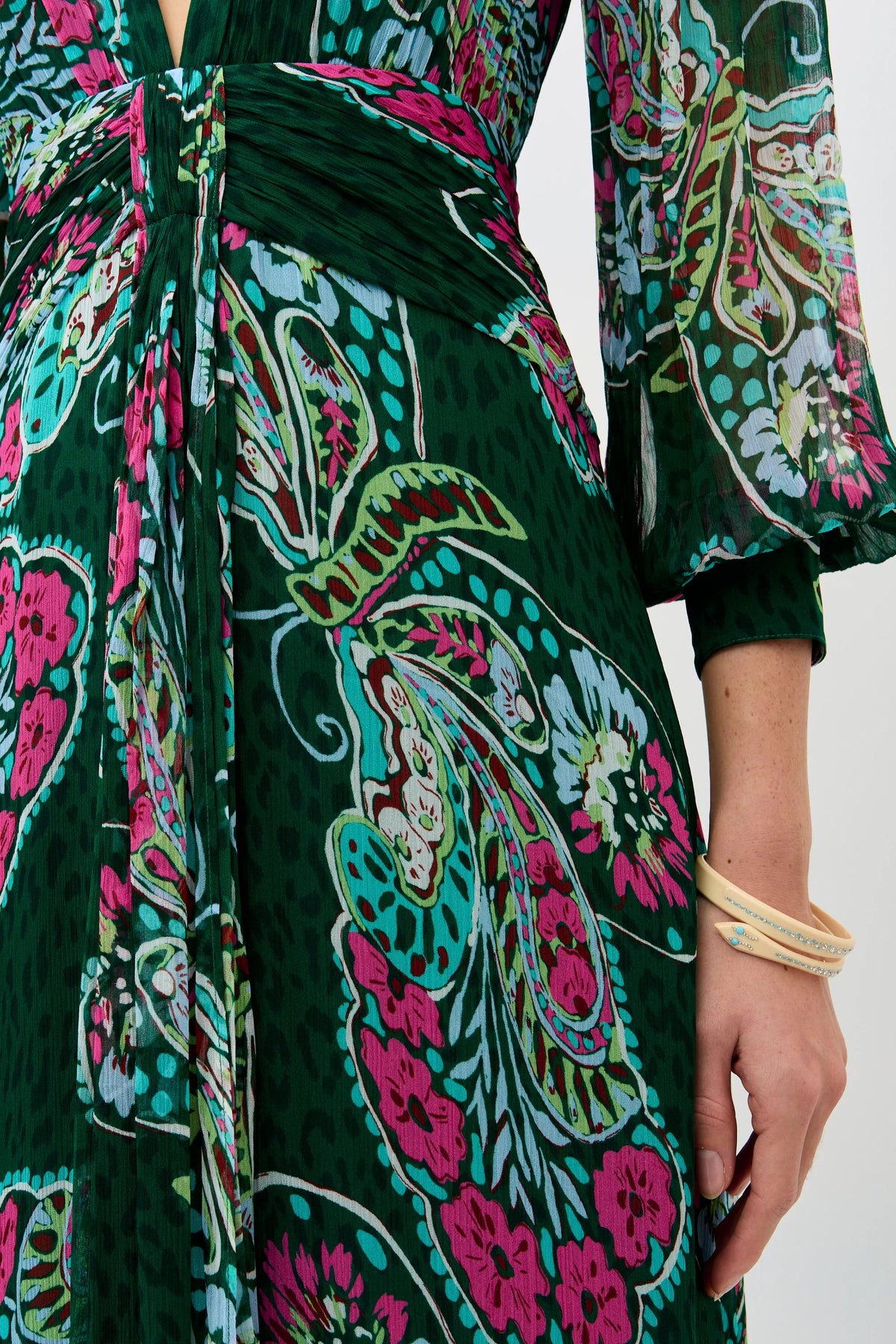 A person wearing the Meera - Butterfly Bloom Green dress, featuring colorful floral and paisley patterns in pink, teal, and white, with light-colored bracelets. The image highlights their midsection and arm.
