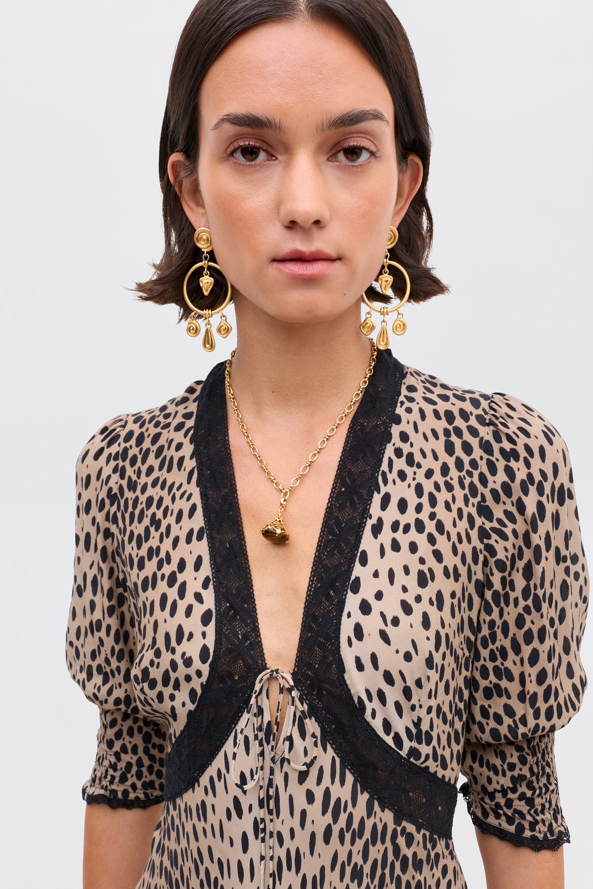 A woman with short brown hair wears statement gold earrings, a gold necklace, and the Amina - Antelope Spot Black dress with elegant black lace trims, standing against a plain light background.