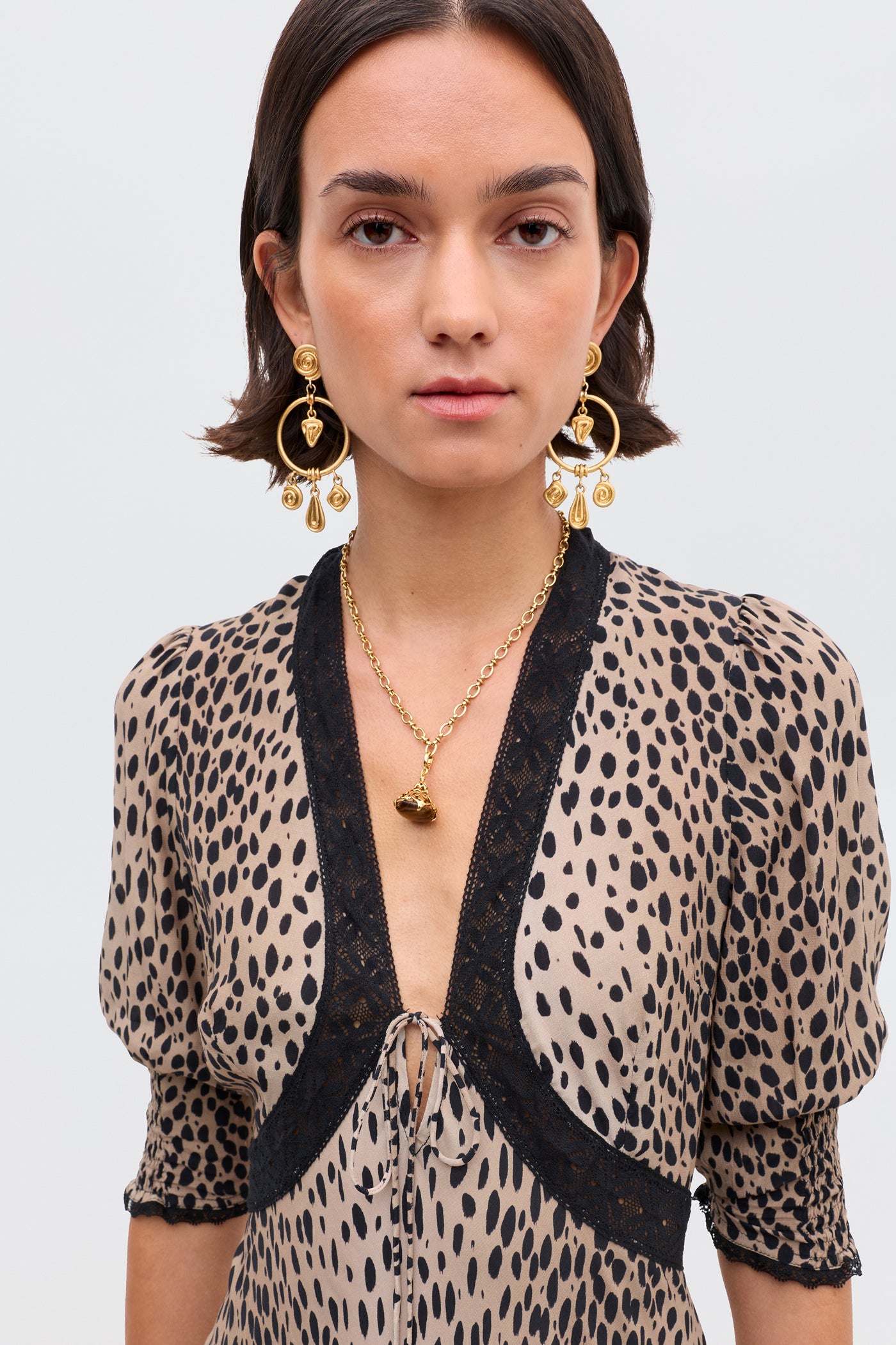 A woman with short brown hair wears statement gold earrings, a gold necklace, and the Amina - Antelope Spot Black dress with elegant black lace trims, standing against a plain light background.