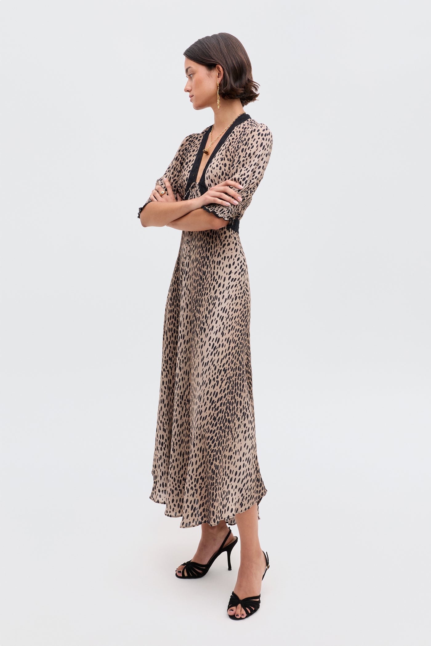 A woman with short dark hair and gold hoop earrings stands arms crossed, wearing the Amina - Antelope Spot Black dress and black high-heeled sandals, looking to the side against a plain light background.