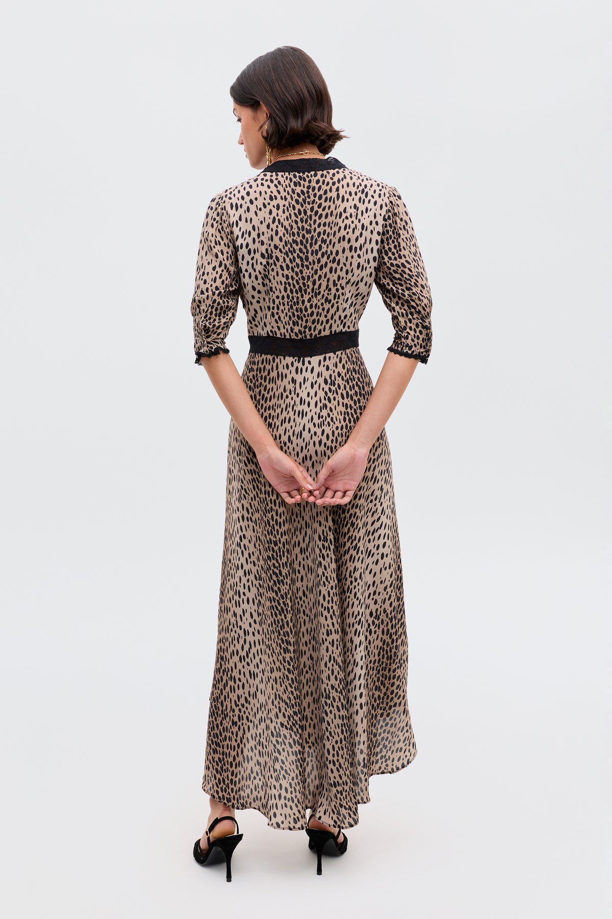 A woman faces away, wearing the Amina - Antelope Spot Black dress—an ankle-length beige style with black spots and accents at the waist, sleeves, and collar. She has short dark hair, black heels, and her hands are clasped behind her back.