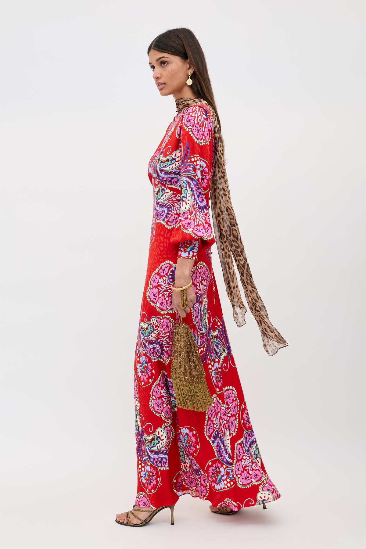 A woman stands in profile wearing the Marni - Butterfly Bloom Jacquard Red dress, heeled sandals, gold earrings, a leopard-print scarf, and holding a woven clutch against a plain white background.