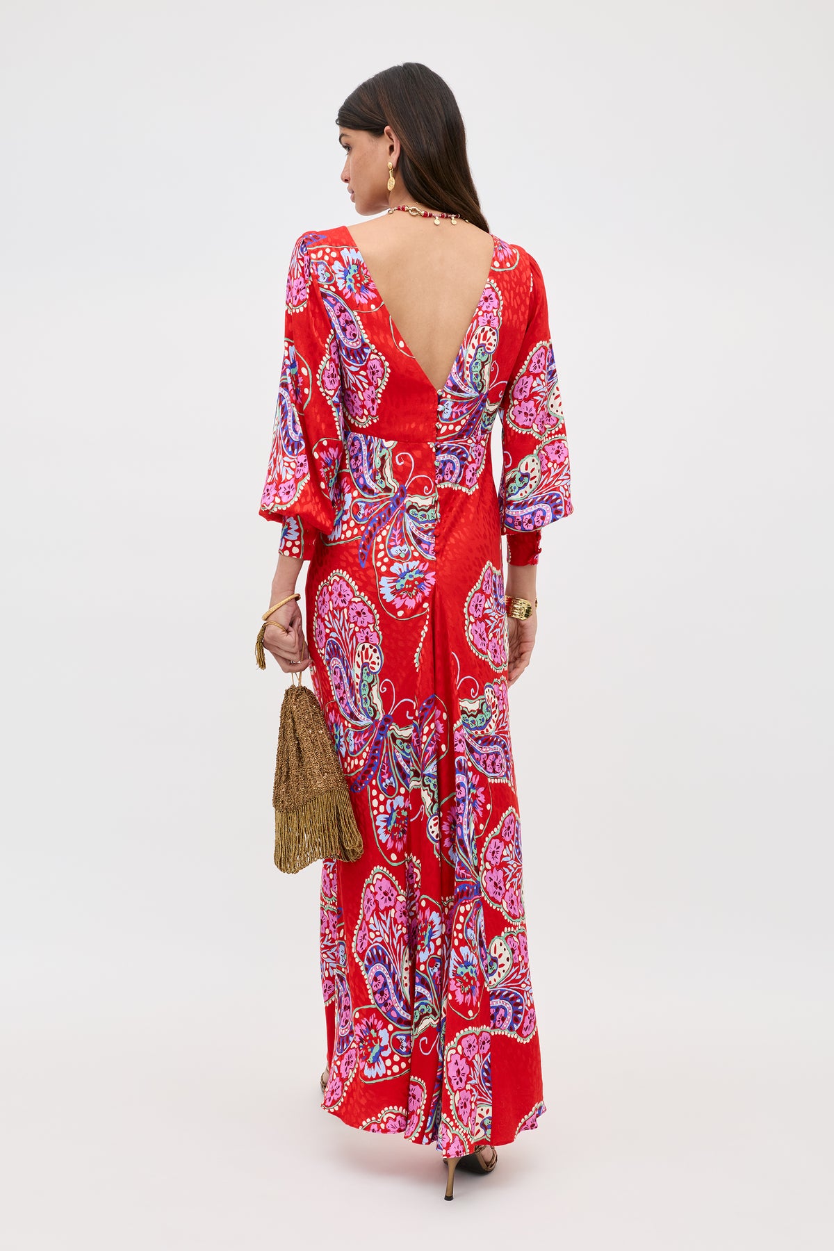 A woman stands facing away, wearing the Marni Butterfly Bloom Jacquard Red maxi dress with a vibrant paisley pattern, V-shaped back, and flared sleeves. She accessorizes with a brown fringed bag, high heels, and jewelry.