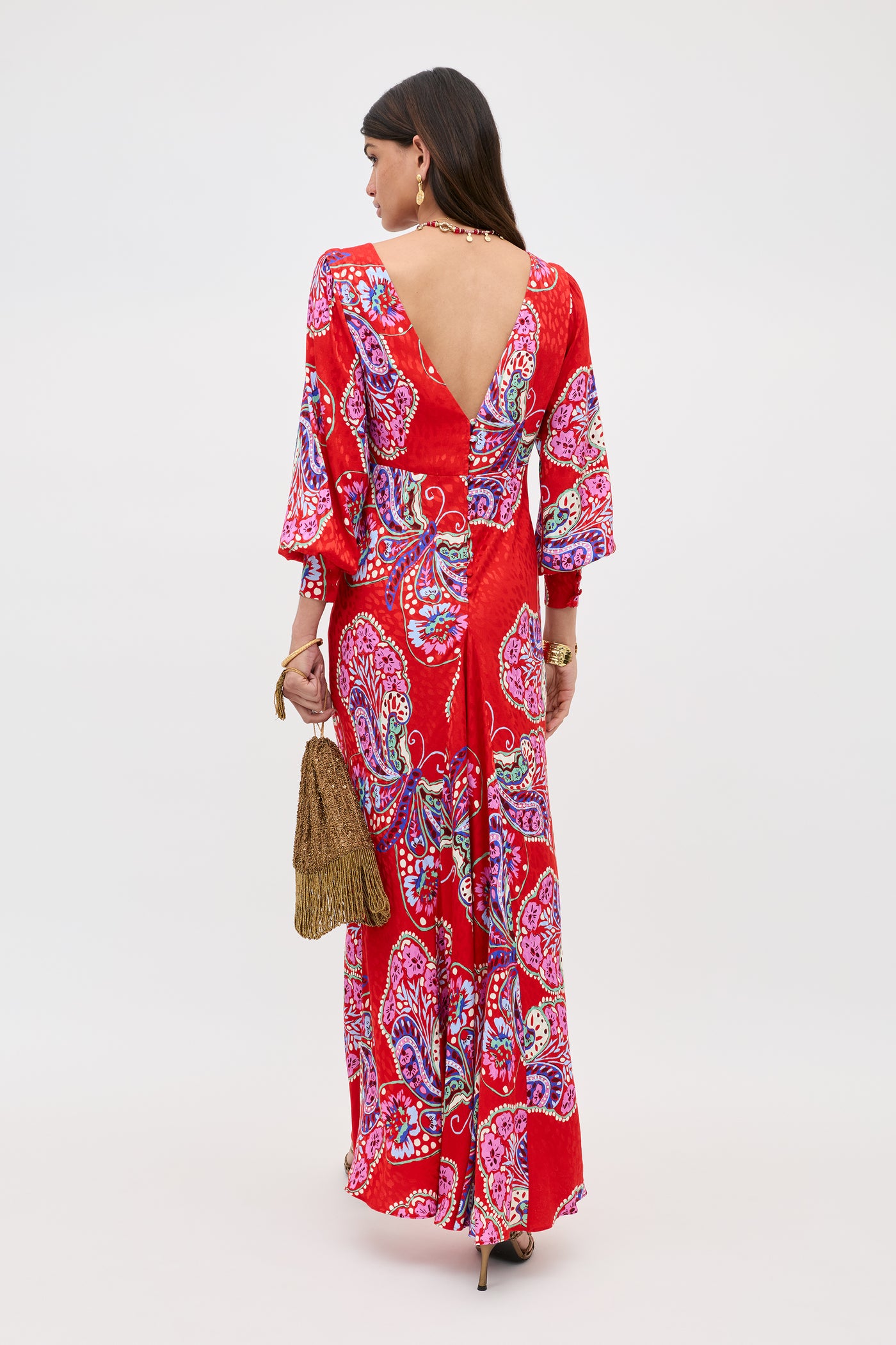 A woman stands facing away, wearing the Marni Butterfly Bloom Jacquard Red maxi dress with a vibrant paisley pattern, V-shaped back, and flared sleeves. She accessorizes with a brown fringed bag, high heels, and jewelry.