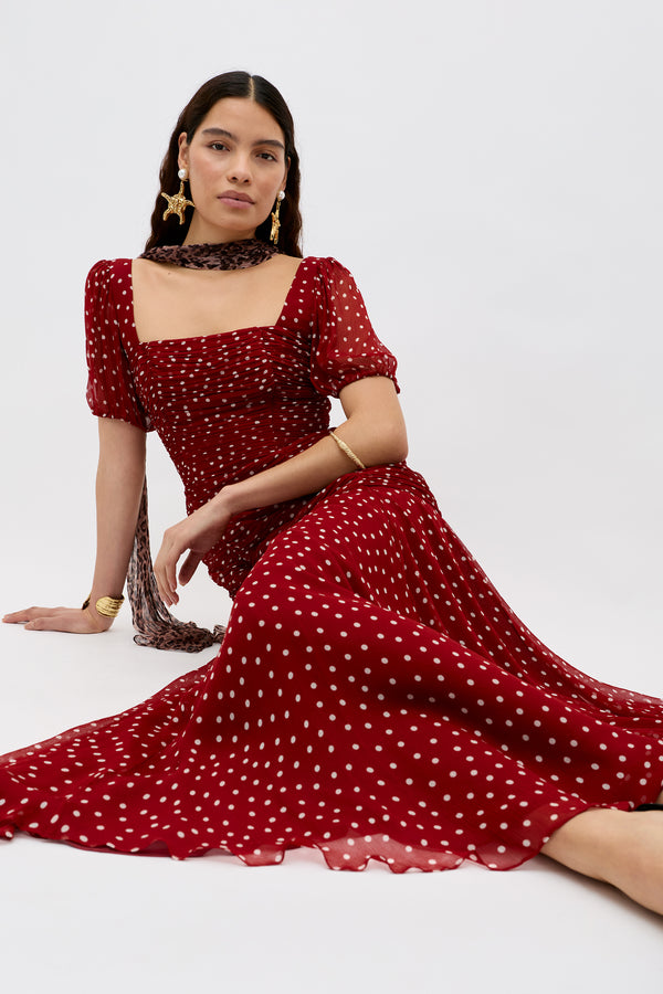 A woman poses sitting on the floor, wearing the Leyane Layla Dot Red dress with white polka dots, puffed short sleeves, and a square neckline. She accessorizes with statement earrings, bracelets, and a scarf.