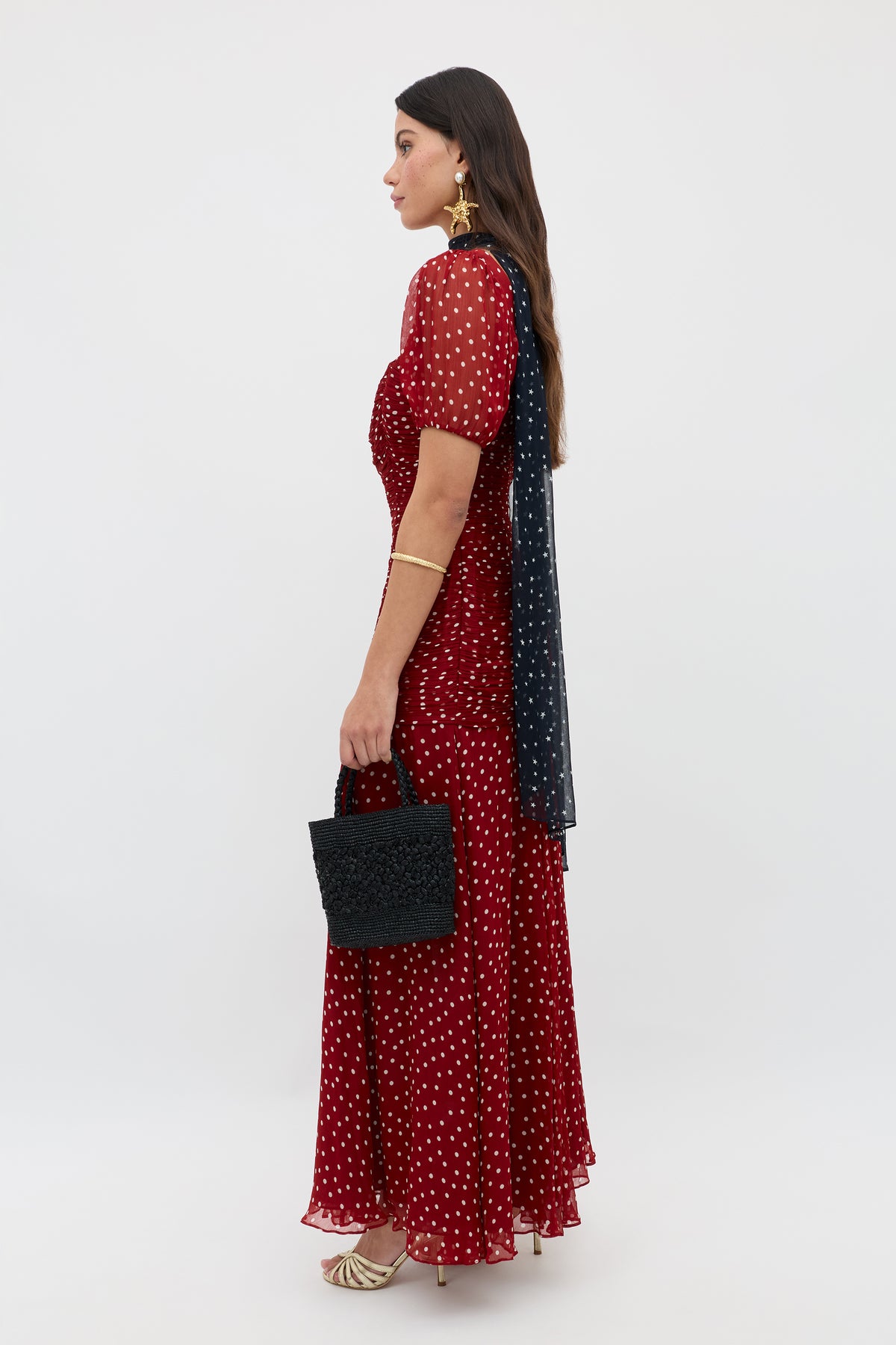 Wearing the Leyane Layla Dot Red dress, a woman stands in profile holding a black textured handbag, styled with gold heels, a gold bracelet, statement earrings, and a dark scarf over one shoulder.