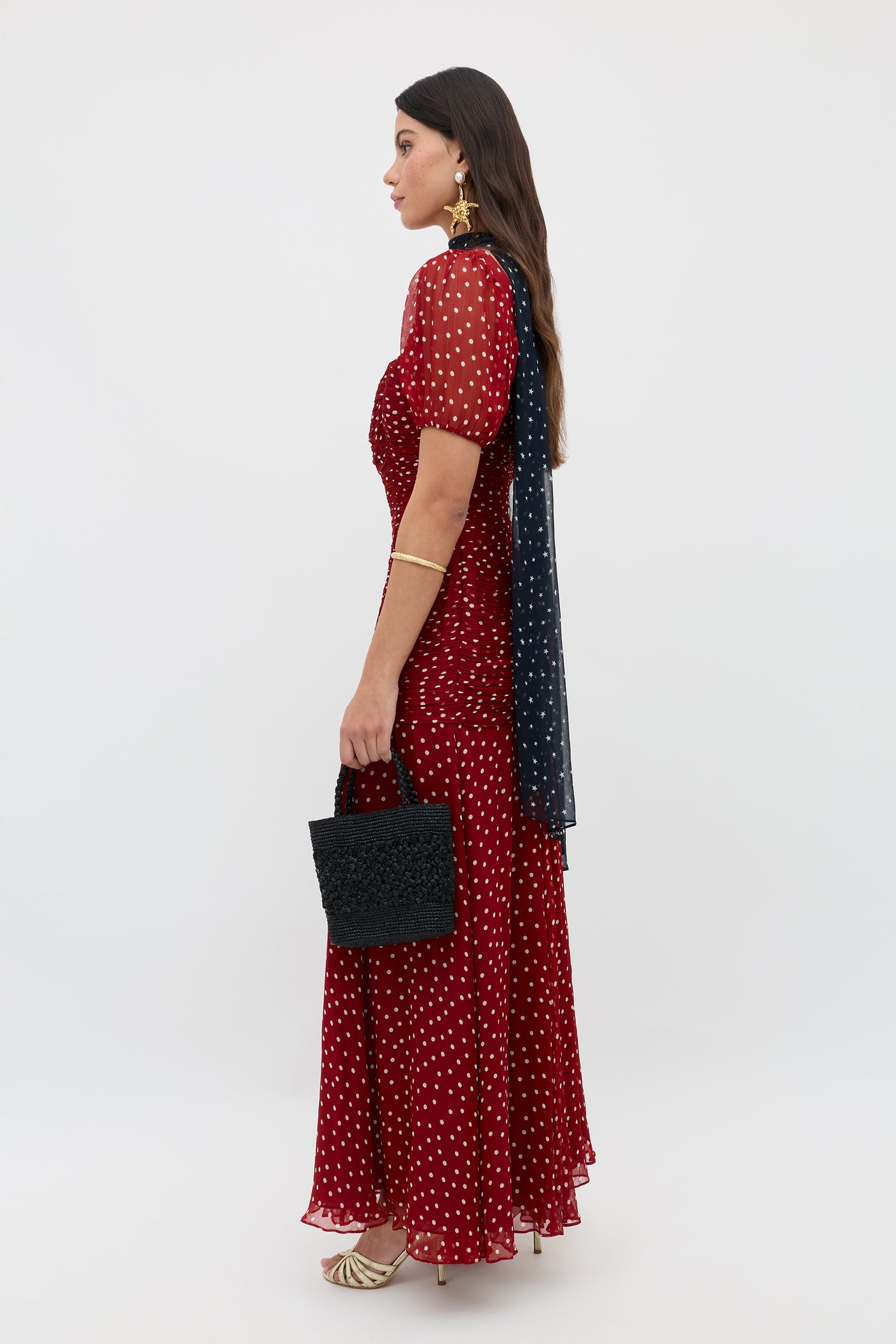 Wearing the Leyane Layla Dot Red dress, a woman stands in profile holding a black textured handbag, styled with gold heels, a gold bracelet, statement earrings, and a dark scarf over one shoulder.