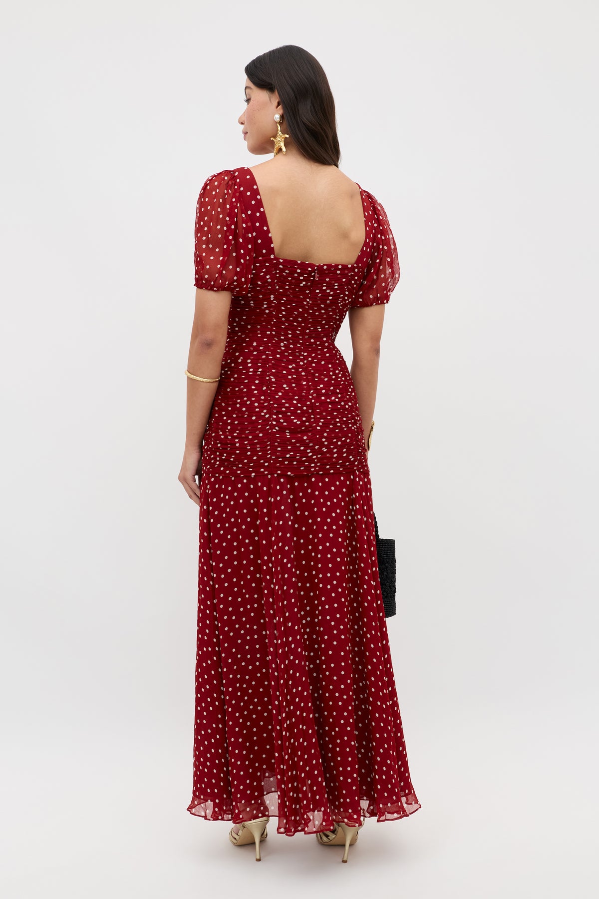 A woman stands facing away, wearing the Leyane Layla Dot Red dress—an ankle-length red dress with white polka dots, puff sleeves, and a square neckline. She accessorizes with a black purse, gold heels, earrings, and a bracelet.