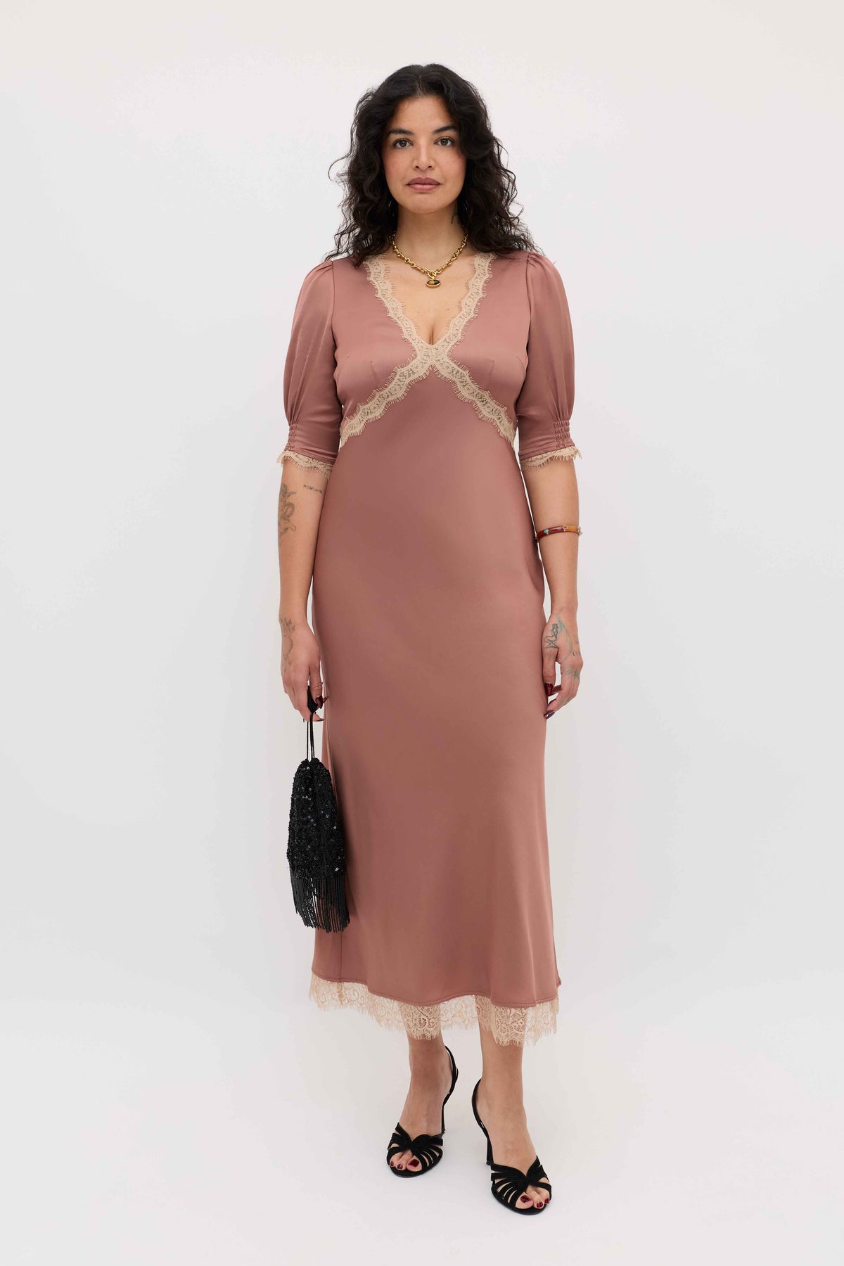 A woman with wavy dark hair and a neutral expression stands against a plain background, wearing the ankle-length Annina - Mocha dress with lace trim, black sandals, gold jewelry, and holding a black handbag.