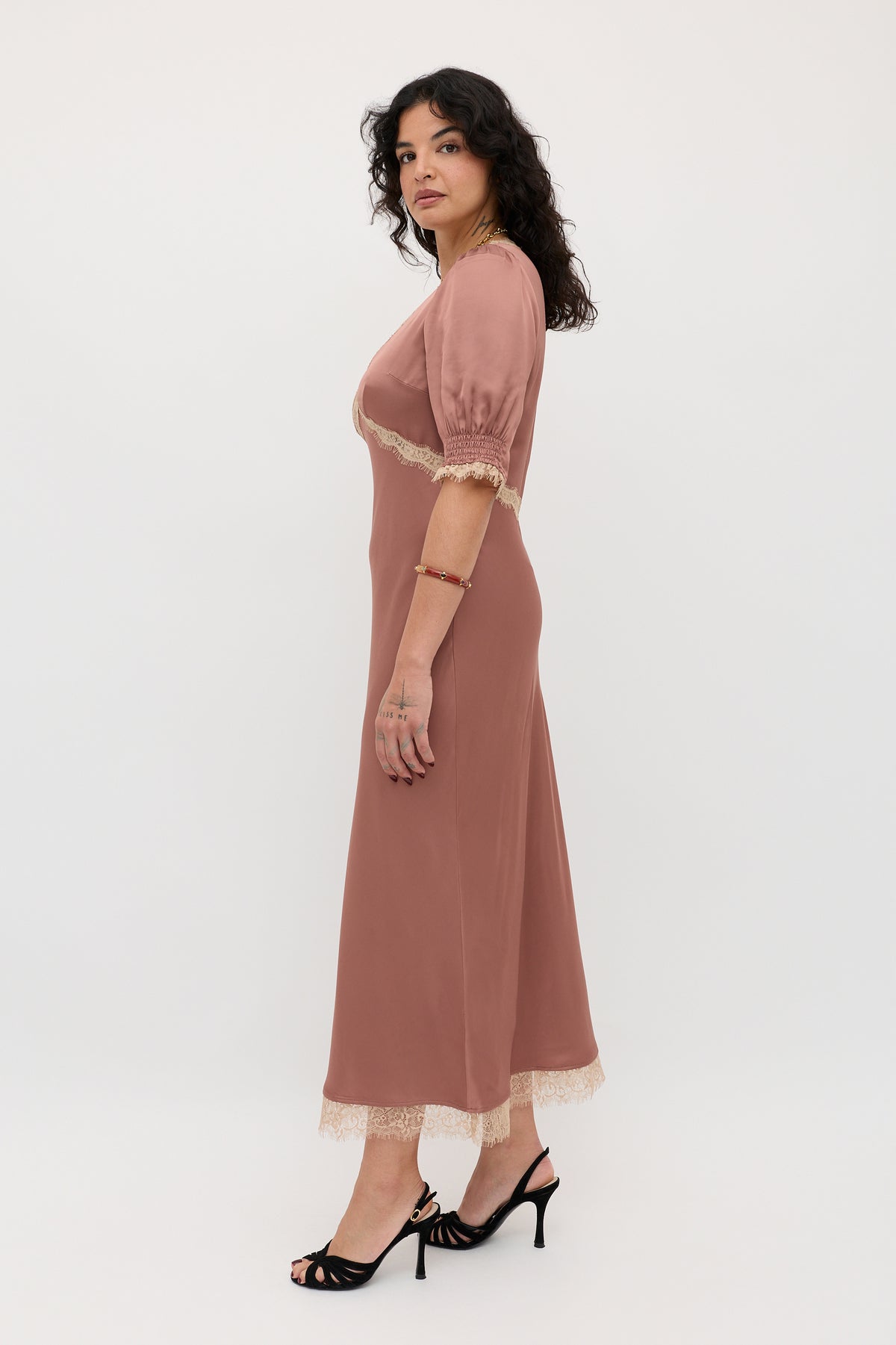 A woman with curly dark hair models the Annina - Mocha, an ankle-length dress with lace trim and short sleeves, paired with black heeled sandals, standing sideways against a plain white background.