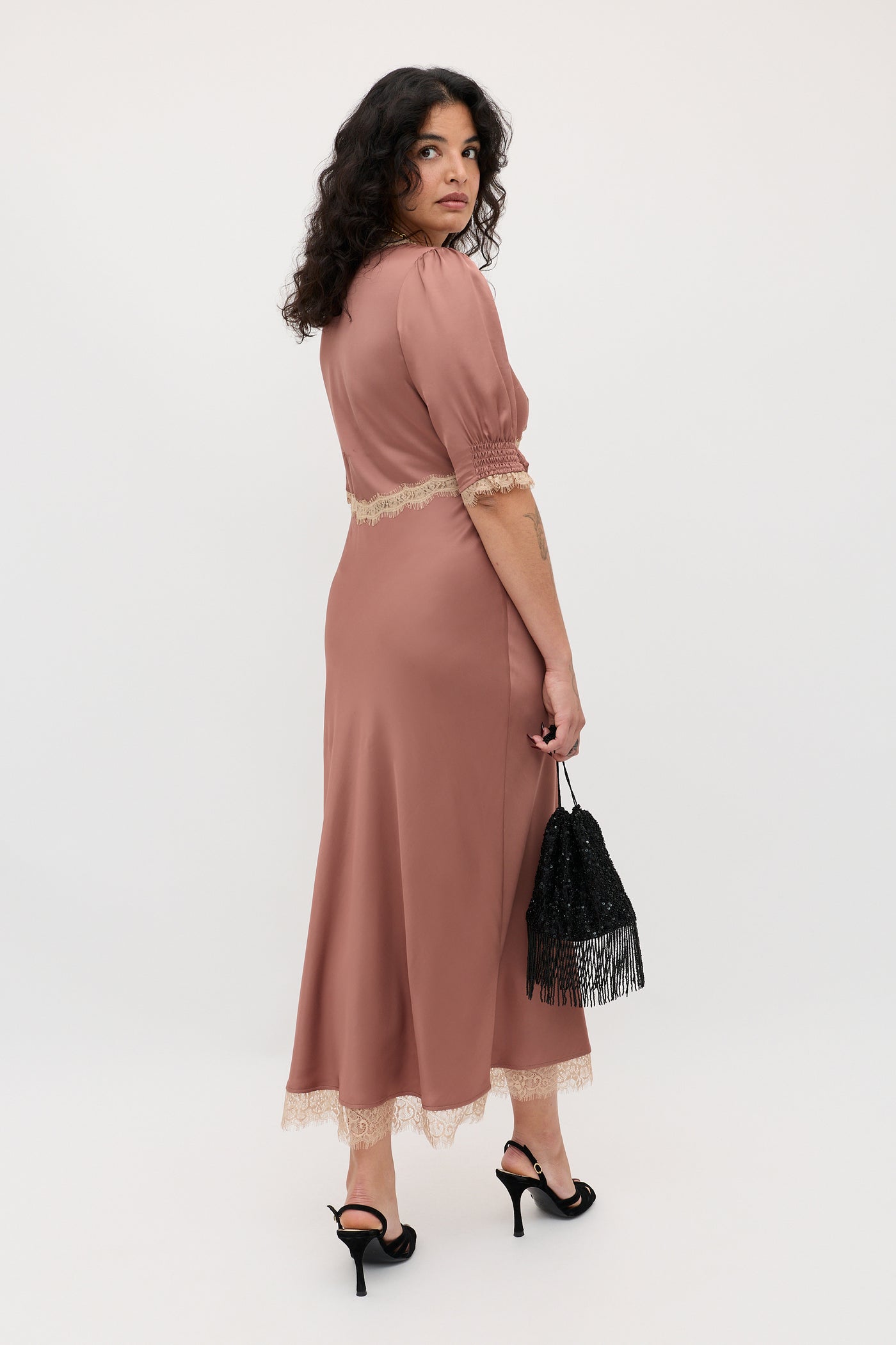 A woman with curly dark hair wears the Annina - Mocha short-sleeved midi dress, black heeled sandals, and holds a black fringe purse while posing over her shoulder against a plain light background.