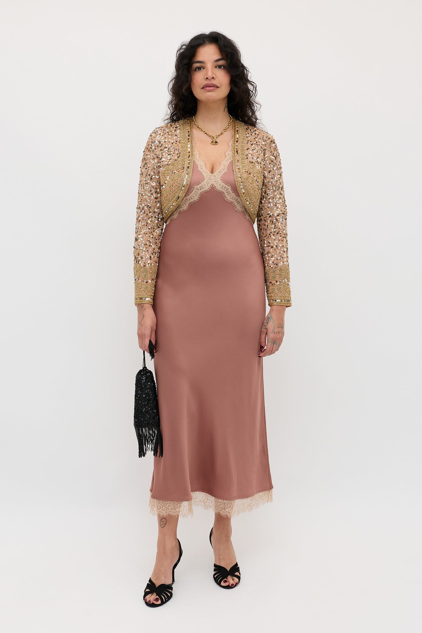 A woman stands against a white background wearing the Annina - Mocha satin slip dress with lace trim, a gold sequin jacket, black strappy heels, layered necklaces, and holding a black fringed clutch.