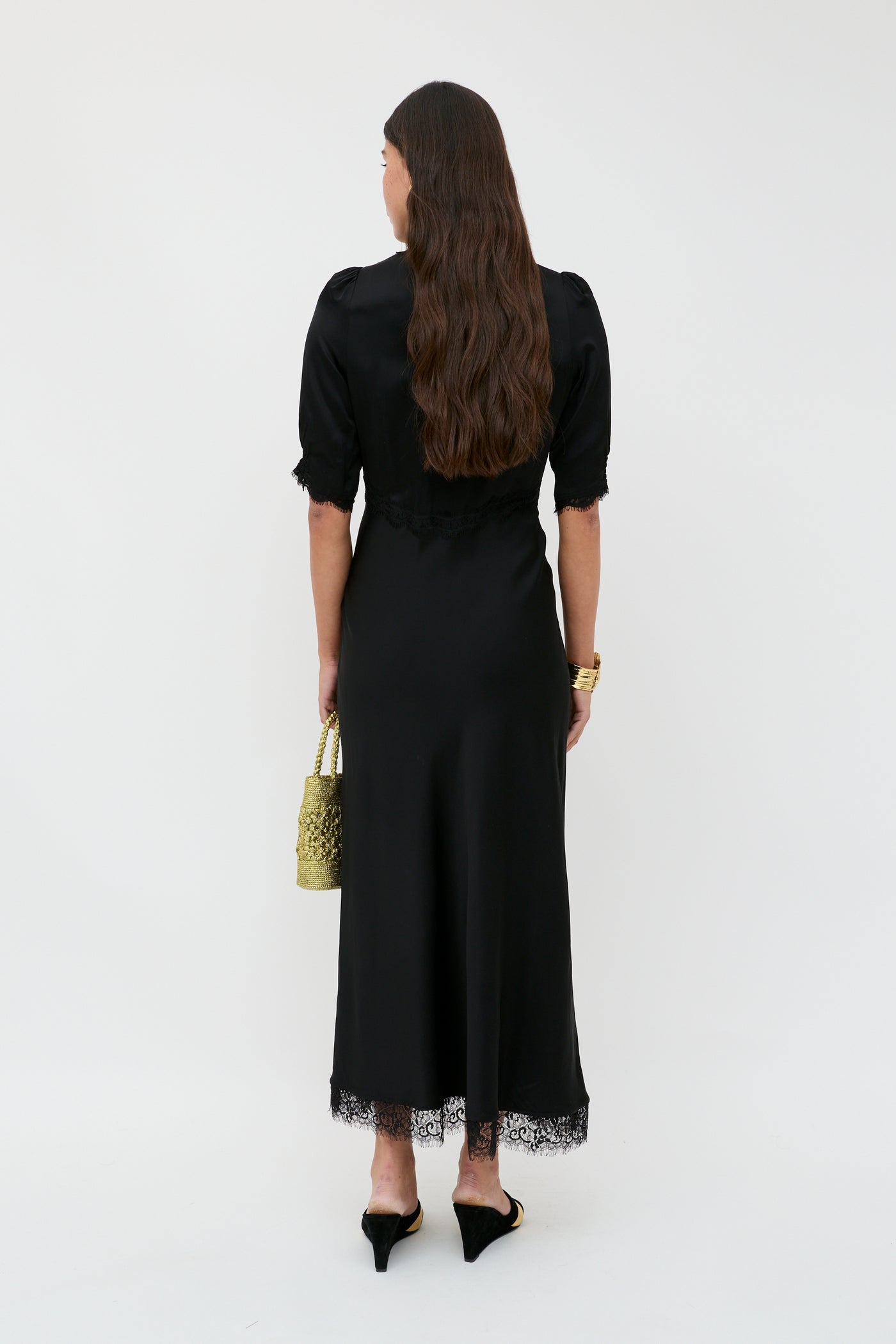A woman with long brown hair stands facing away, wearing the Annina - Black midi dress with lace trims and black heels, holding a yellow textured handbag. The bias cut flatters her silhouette against a plain white background.