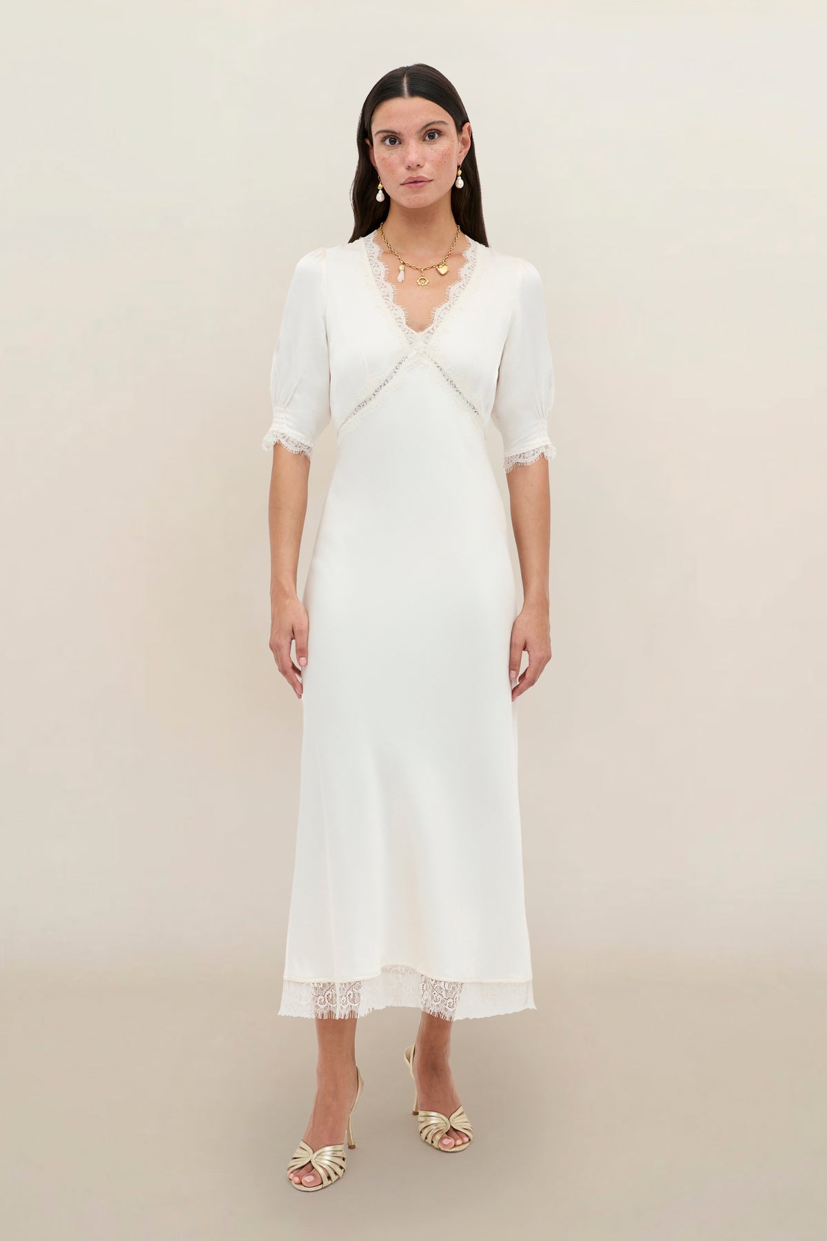 A woman with long dark hair stands against a plain light background, wearing the Annina - Pearl dress—a white, mid-length vintage style with lace trim and puff half sleeves—paired with cream heeled sandals and gold jewelry.