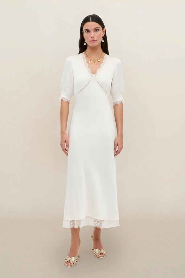 A woman with long dark hair stands against a plain light background, wearing the Annina - Pearl dress—a white, mid-length vintage style with lace trim and puff half sleeves—paired with cream heeled sandals and gold jewelry.