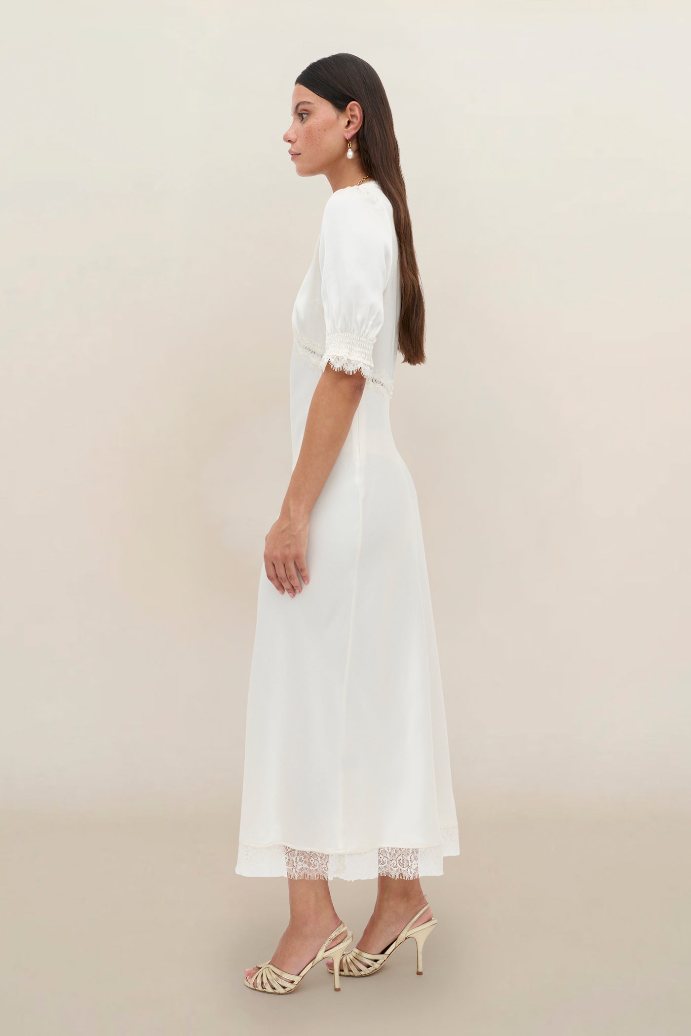 A woman stands in profile against a plain background, wearing the Annina - Pearl, a white mid-length vintage dress with lace trim on the sleeves and hem, paired with open-toe heels.