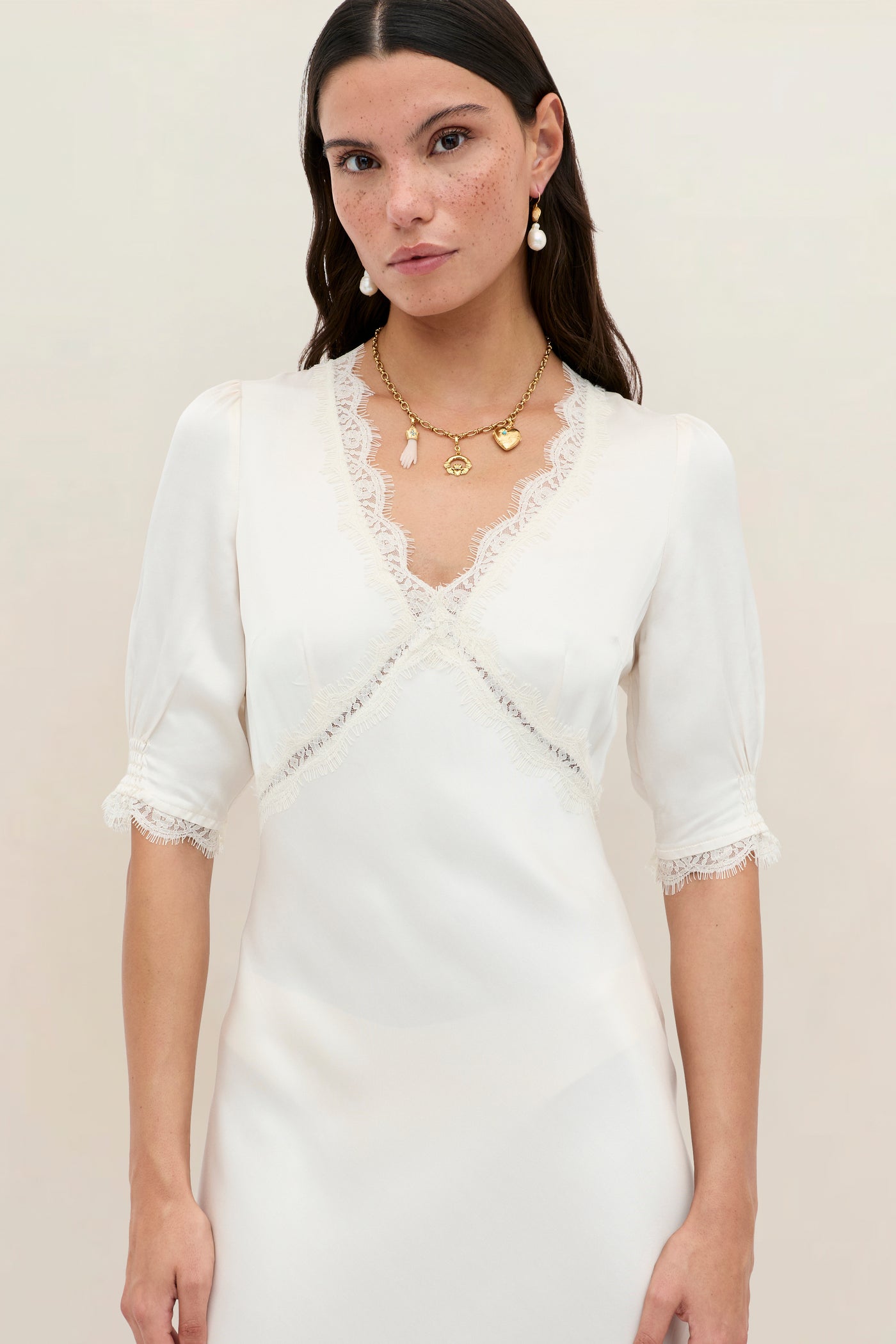 A woman in the Annina - Pearl dress, a vintage style with lace trim, short sleeves, and a V-neckline. She has long brown hair, drop earrings, and a gold pendant necklace against a plain light-colored background.