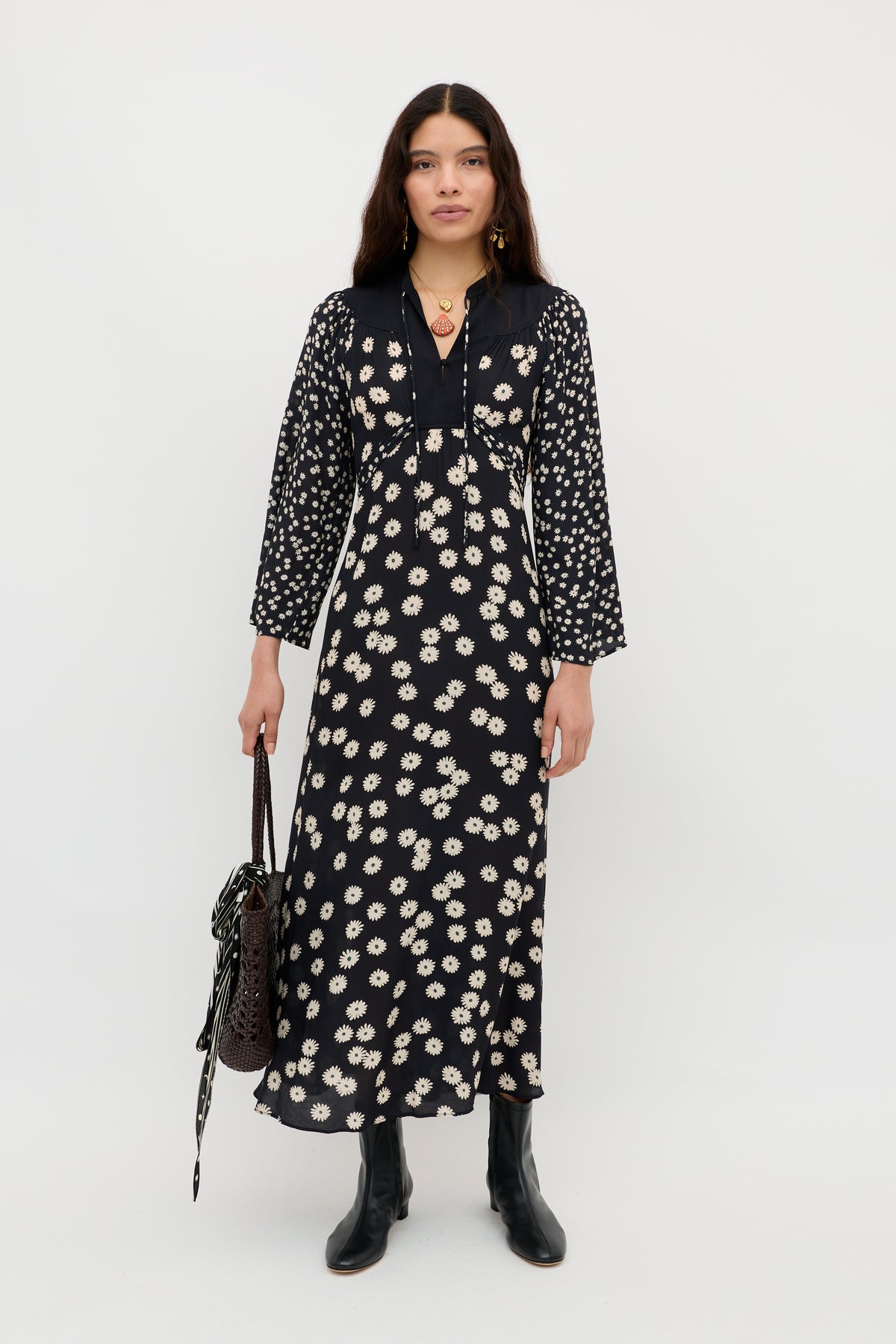 A woman stands against a plain white background wearing the Anela - Daisy Mix Black long boho dress with a white floral pattern, black boots, and carrying a textured dark handbag—perfect for stylish everyday wear.