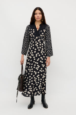 A woman stands against a plain white background wearing the Anela - Daisy Mix Black long boho dress with a white floral pattern, black boots, and carrying a textured dark handbag—perfect for stylish everyday wear.