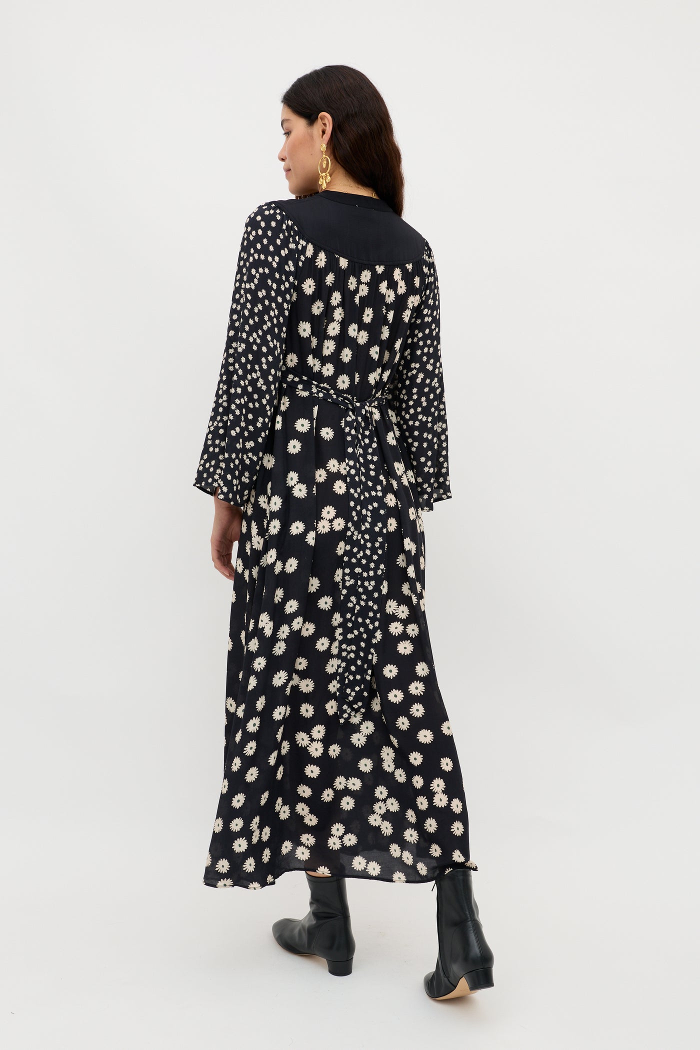 A woman with long dark hair wears the Anela - Daisy Mix Black dress, an ankle-length boho style featuring a white floral pattern, long sleeves, and a waist tie. She stands facing away in black heeled ankle boots and large gold earrings.