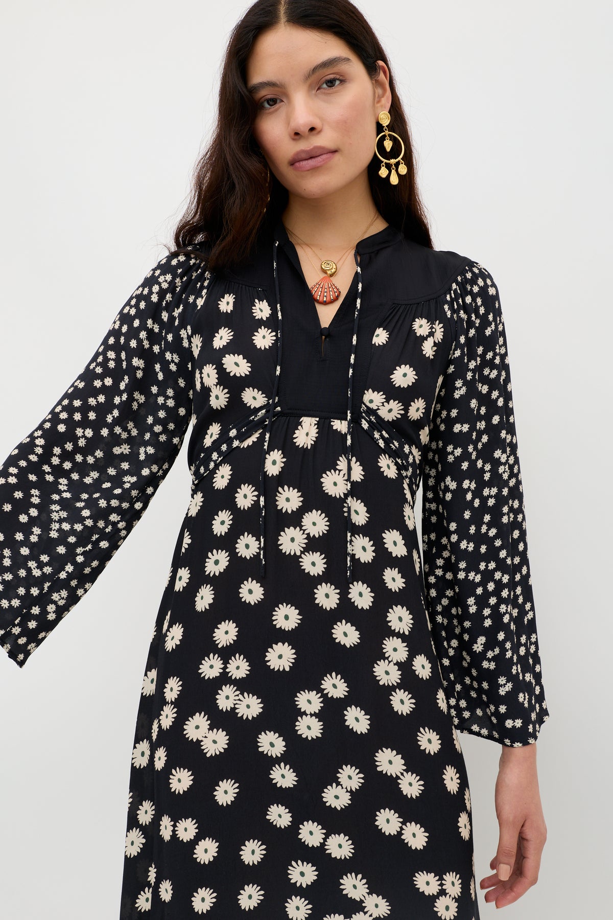 A woman with long dark hair wears the Anela - Daisy Mix Black dress, a lightweight crepe boho piece with white daisy print, long sleeves, and black panel top. She accessorizes with gold earrings and a pendant necklace against a plain white background.