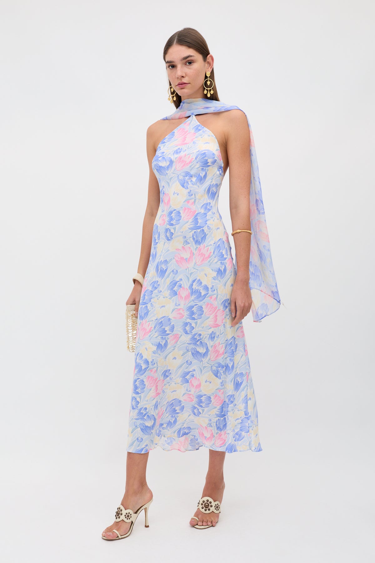 A woman wearing the Hayley - Tulip Fields Blue dress, a sleeveless ankle-length pastel blue and pink floral silk crepe de chine, with white heels, gold earrings, a matching detachable georgette scarf over one shoulder, and a small beaded clutch.