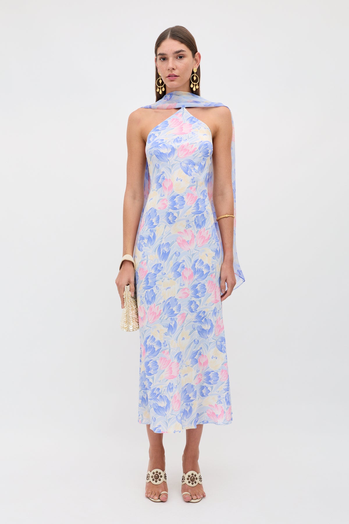 A woman stands against a plain background wearing the Hayley - Tulip Fields Blue dress, a pastel floral halter-neck style in silk crepe de chine with a detachable georgette scarf, styled with gold statement earrings, bracelet, heeled sandals, and a small beaded bag.