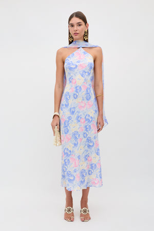 A woman stands against a plain background wearing the Hayley - Tulip Fields Blue dress, a pastel floral halter-neck style in silk crepe de chine with a detachable georgette scarf, styled with gold statement earrings, bracelet, heeled sandals, and a small beaded bag.